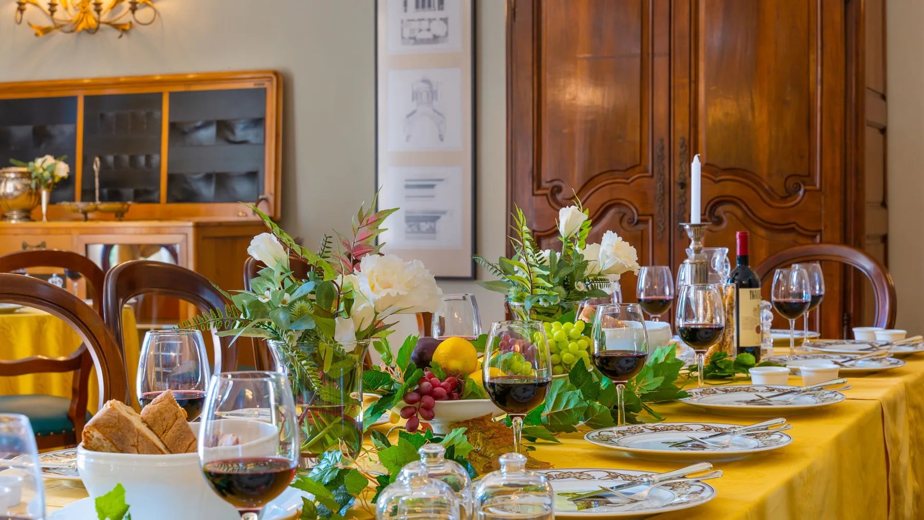 Una mesa de comedor está puesta con platos, copas de vino, pan y arreglos florales.