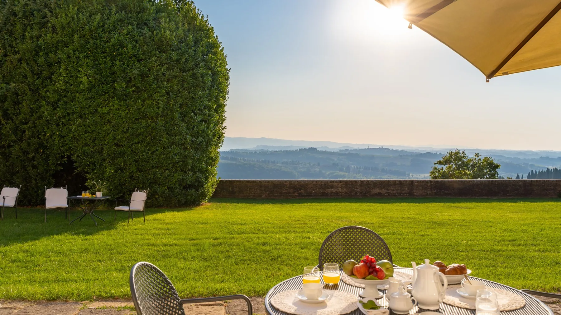 Un jardín con césped y una mesa redonda preparada para comer, con colinas lejanas.