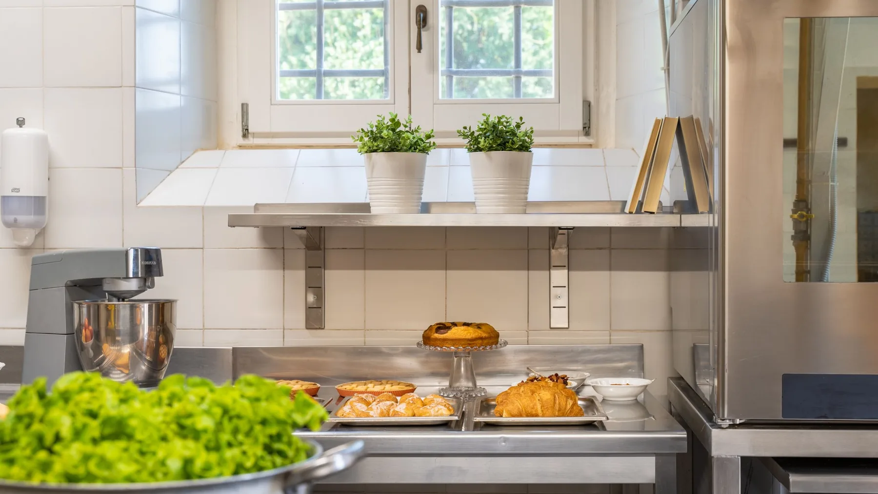 Una encimera de cocina con azulejos exhibe bandejas de productos horneados bajo dos ventanas.