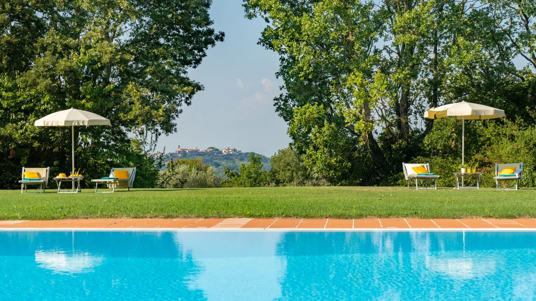 Una piscina rodeada de césped y árboles, con asientos reclinables y sombrillas.