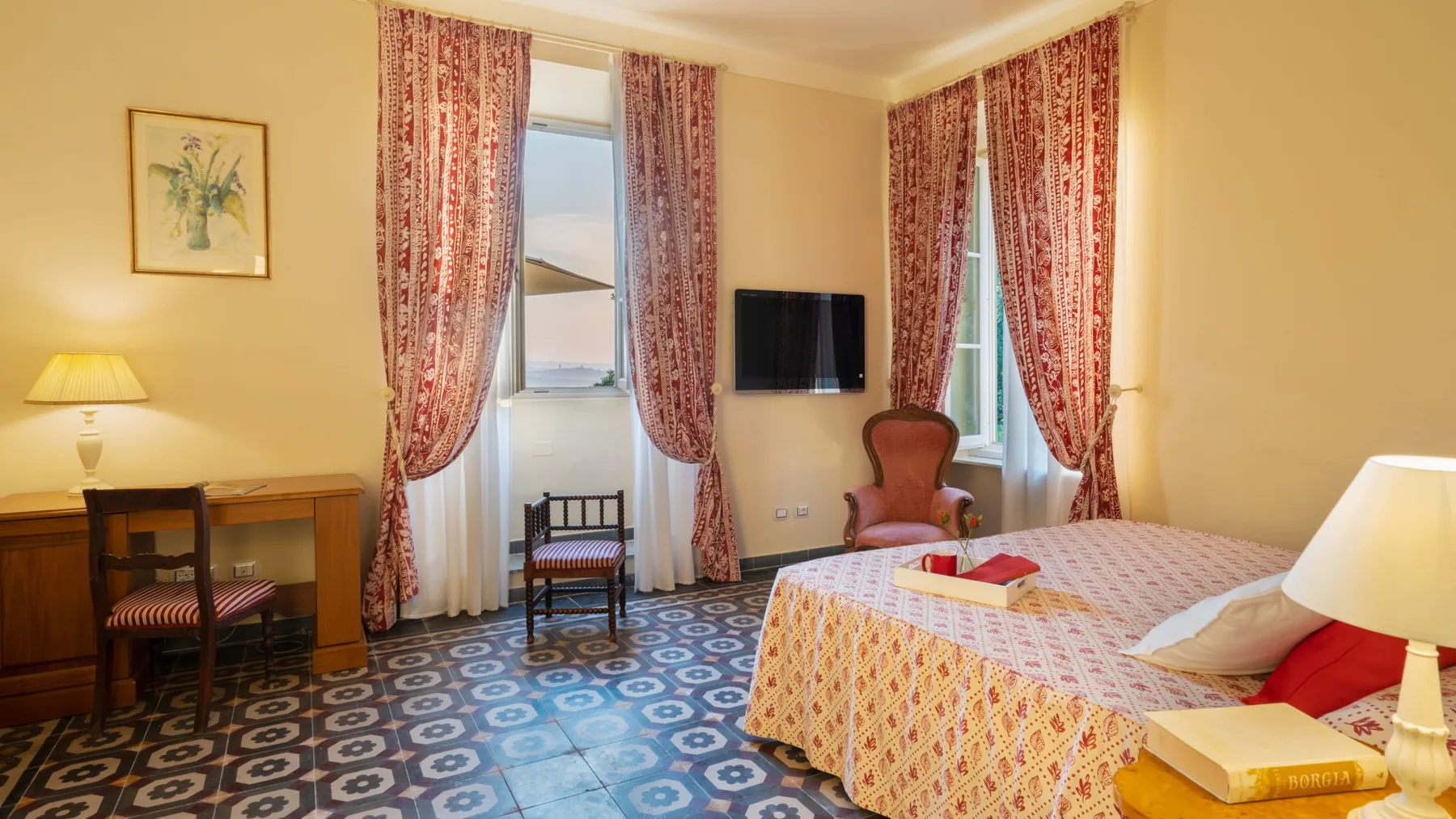 Dormitorio con una colcha estampada, un televisor y ventanas altas con cortinas.