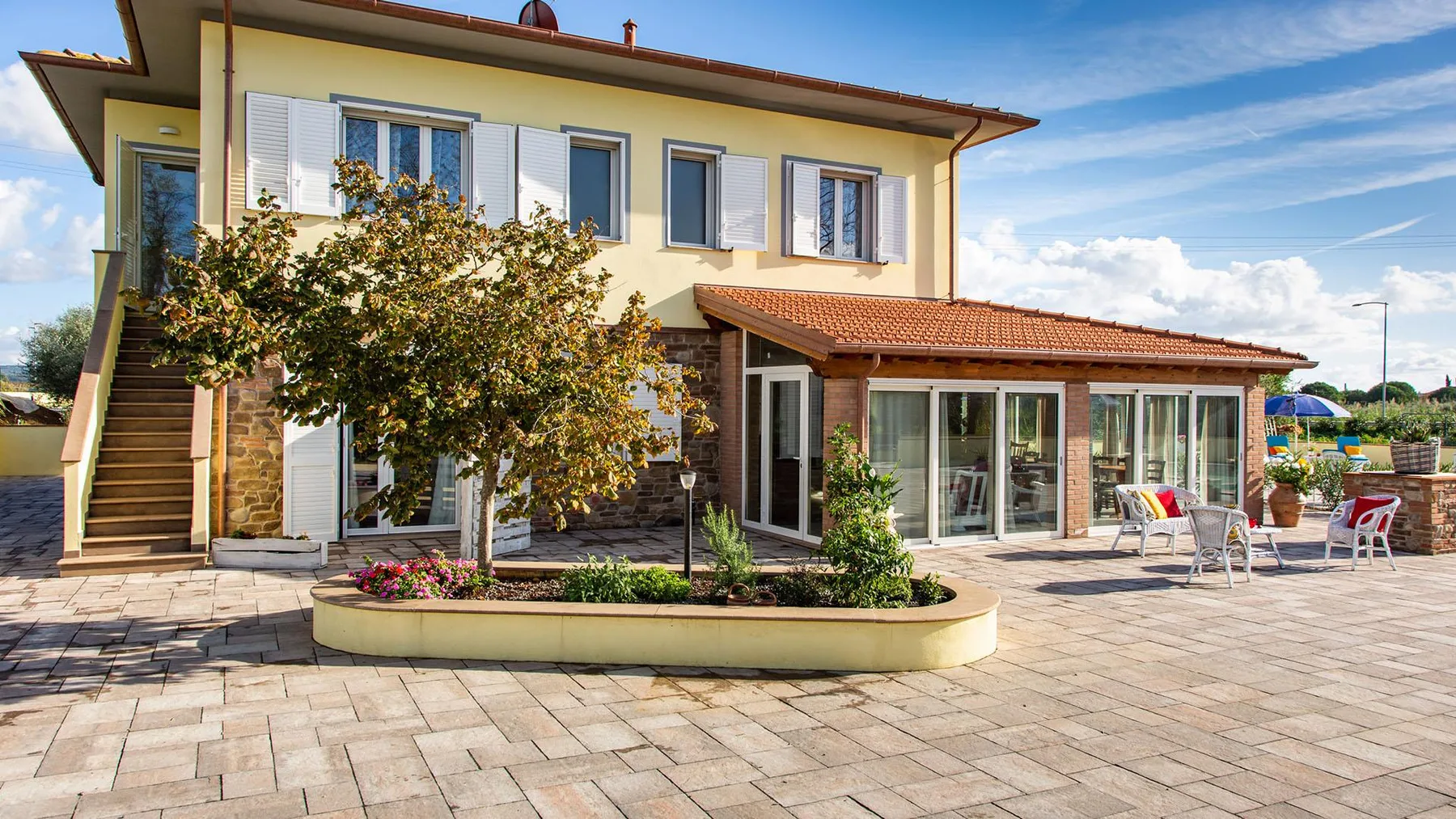 A two-story house opens onto a paved patio with outdoor seating.