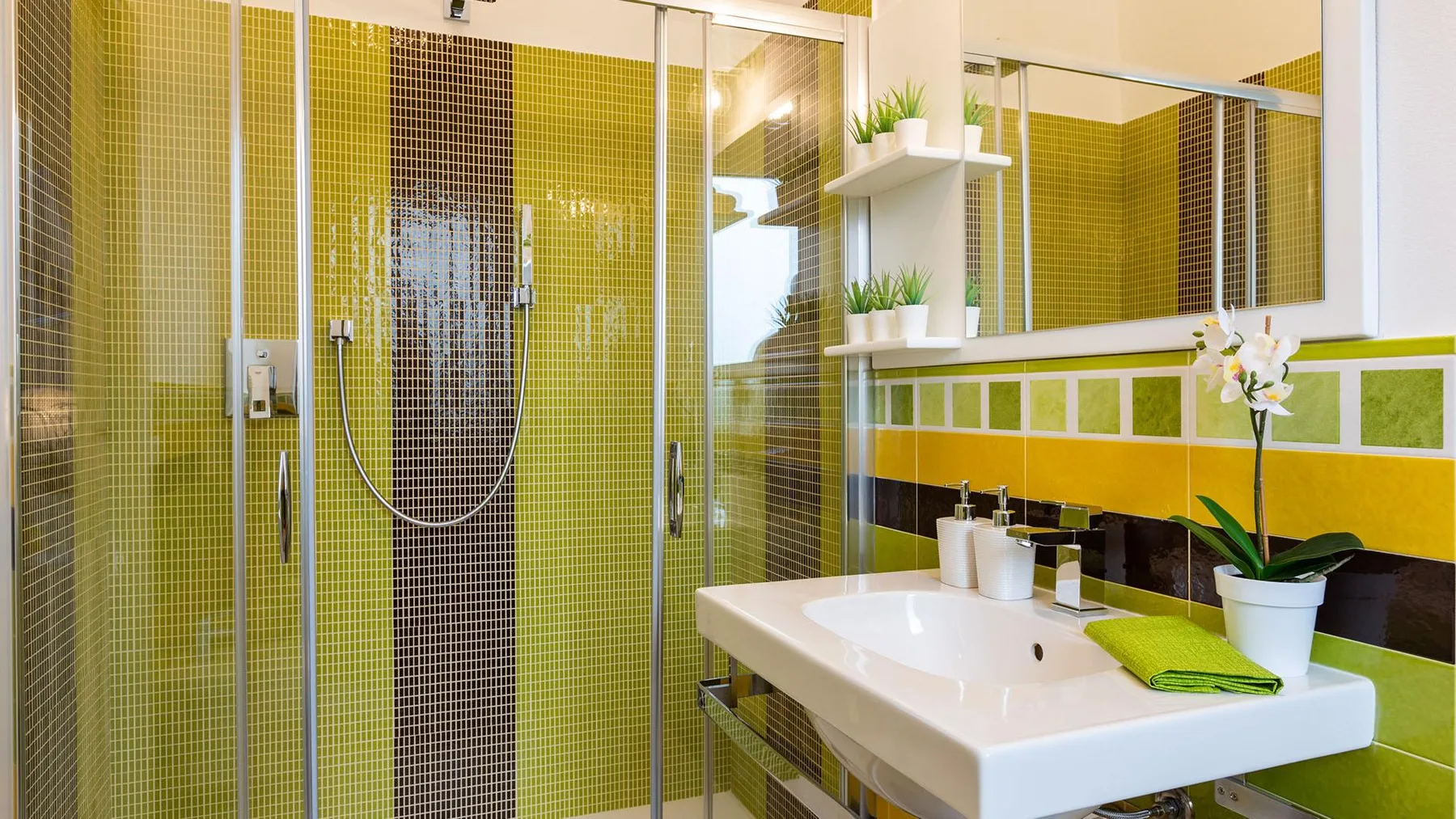 A bright bathroom with a glass shower, sink, and green mosaic tile.