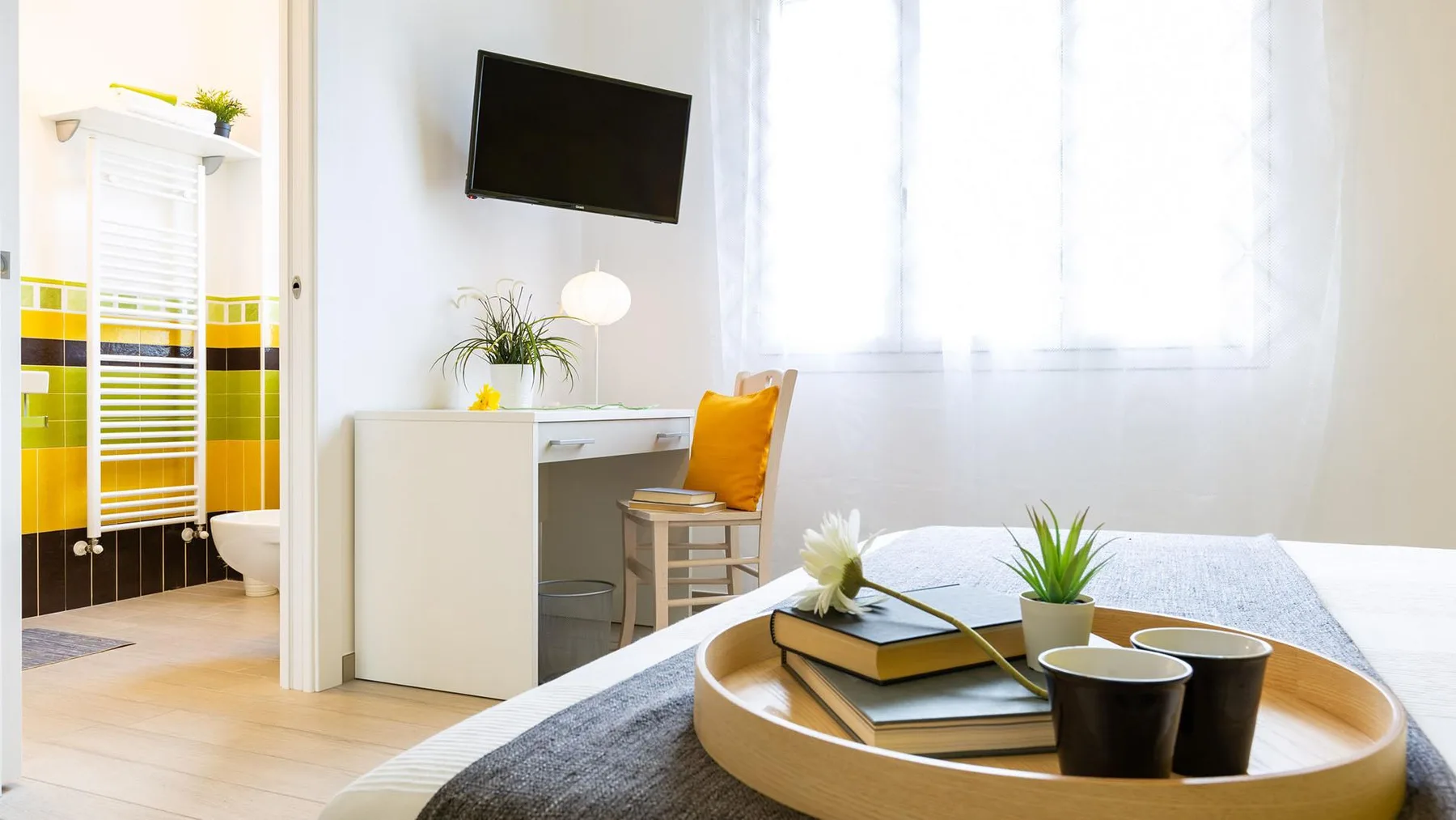 A bright bedroom with a desk, seating, and a round tray on the bed.