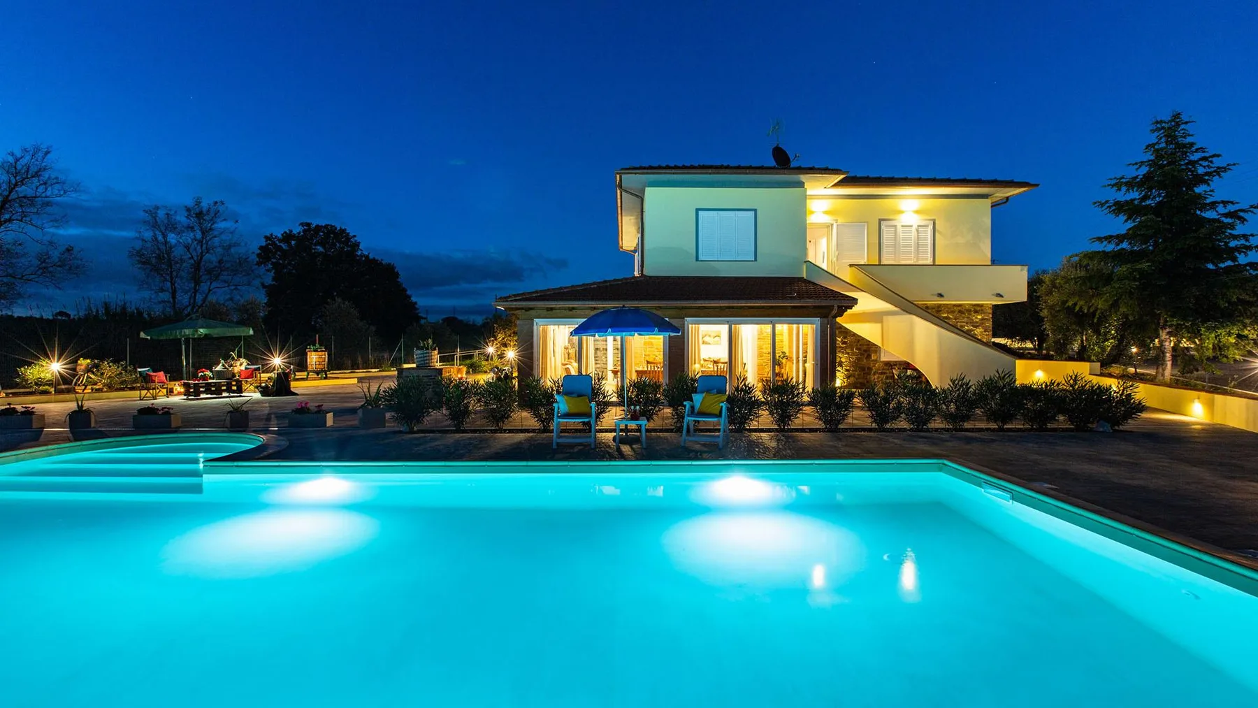 A lit modern house overlooks a swimming pool at dusk.