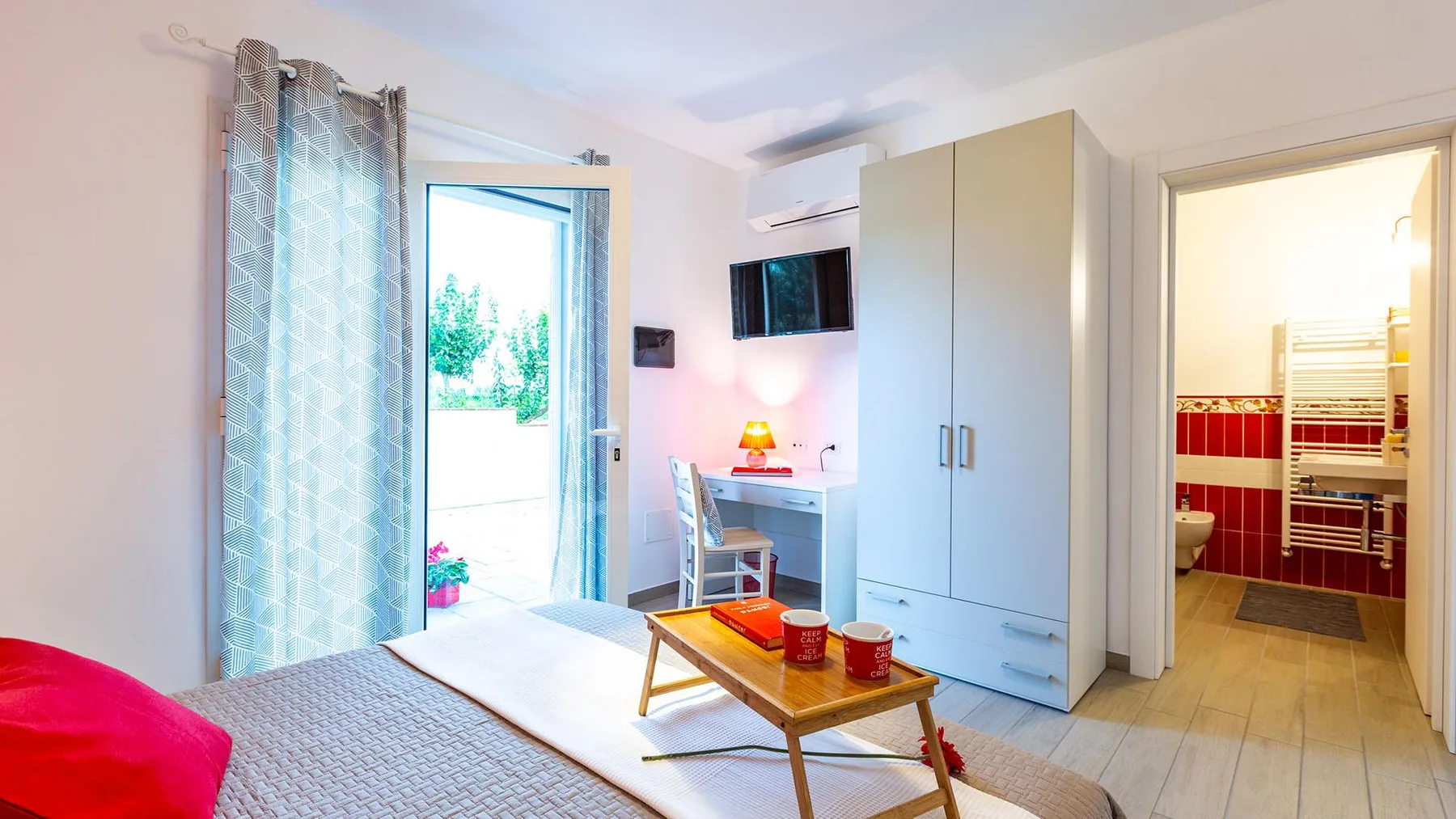 A bright bedroom with a bed, a small table, and an open door to the bathroom.