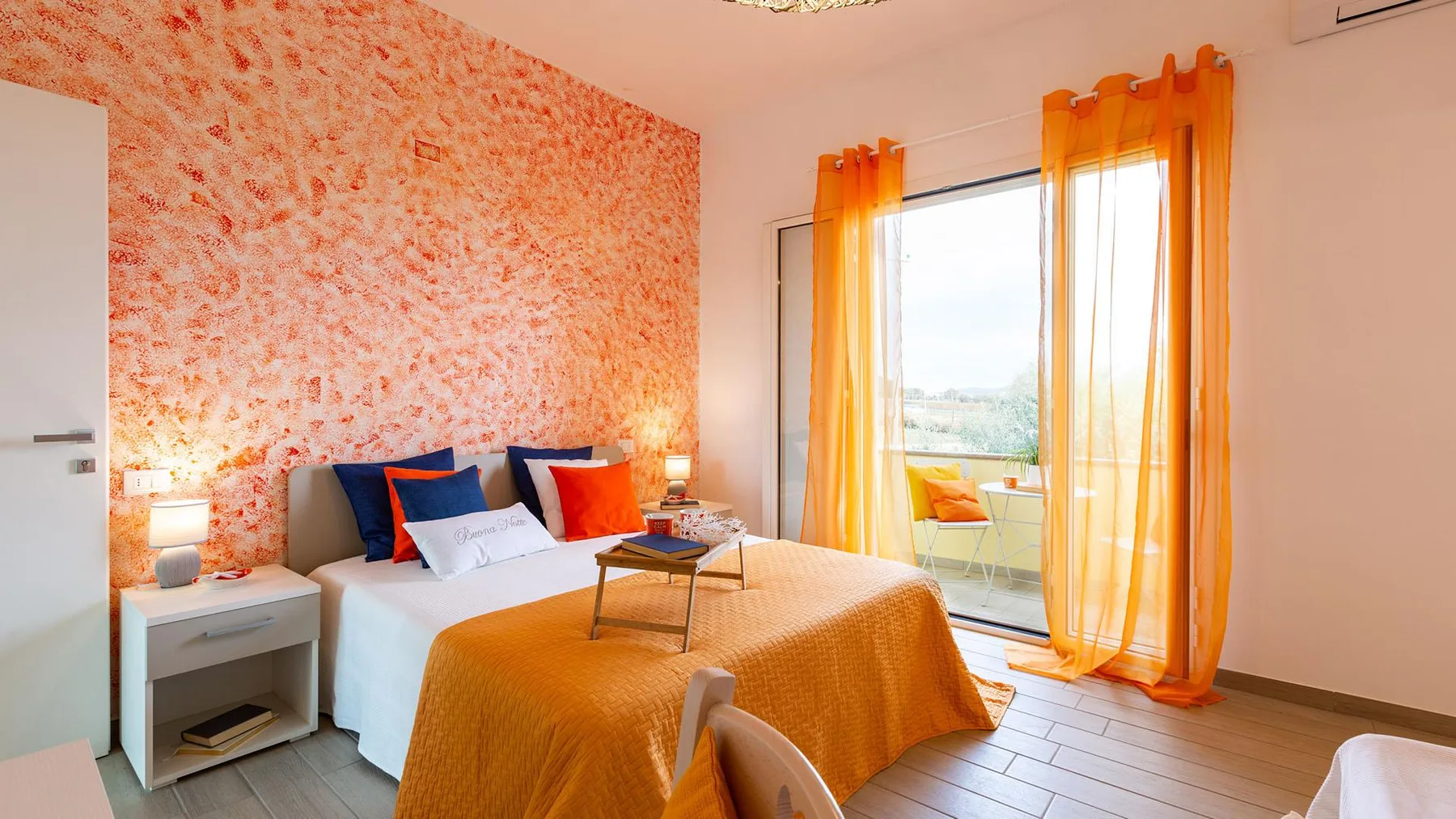 A bedroom with a bed covered by an orange bedspread beside a window.