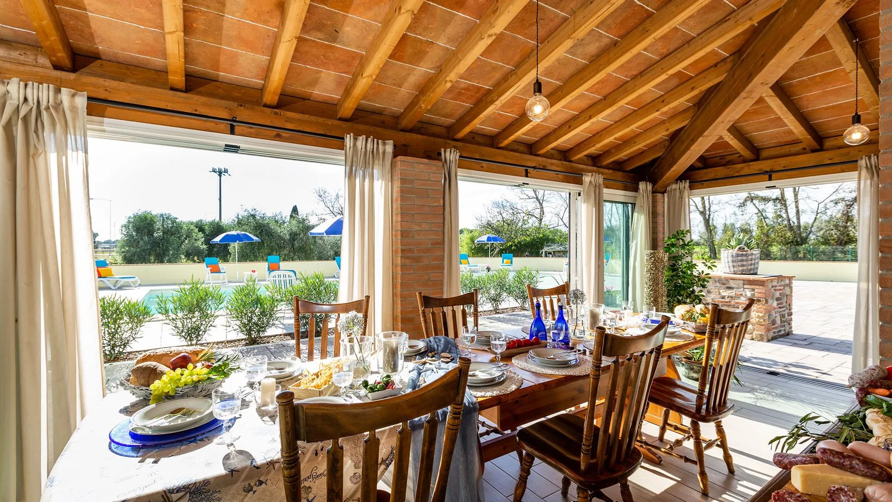 A covered dining area with a long table set beside large windows overlooking a pool.