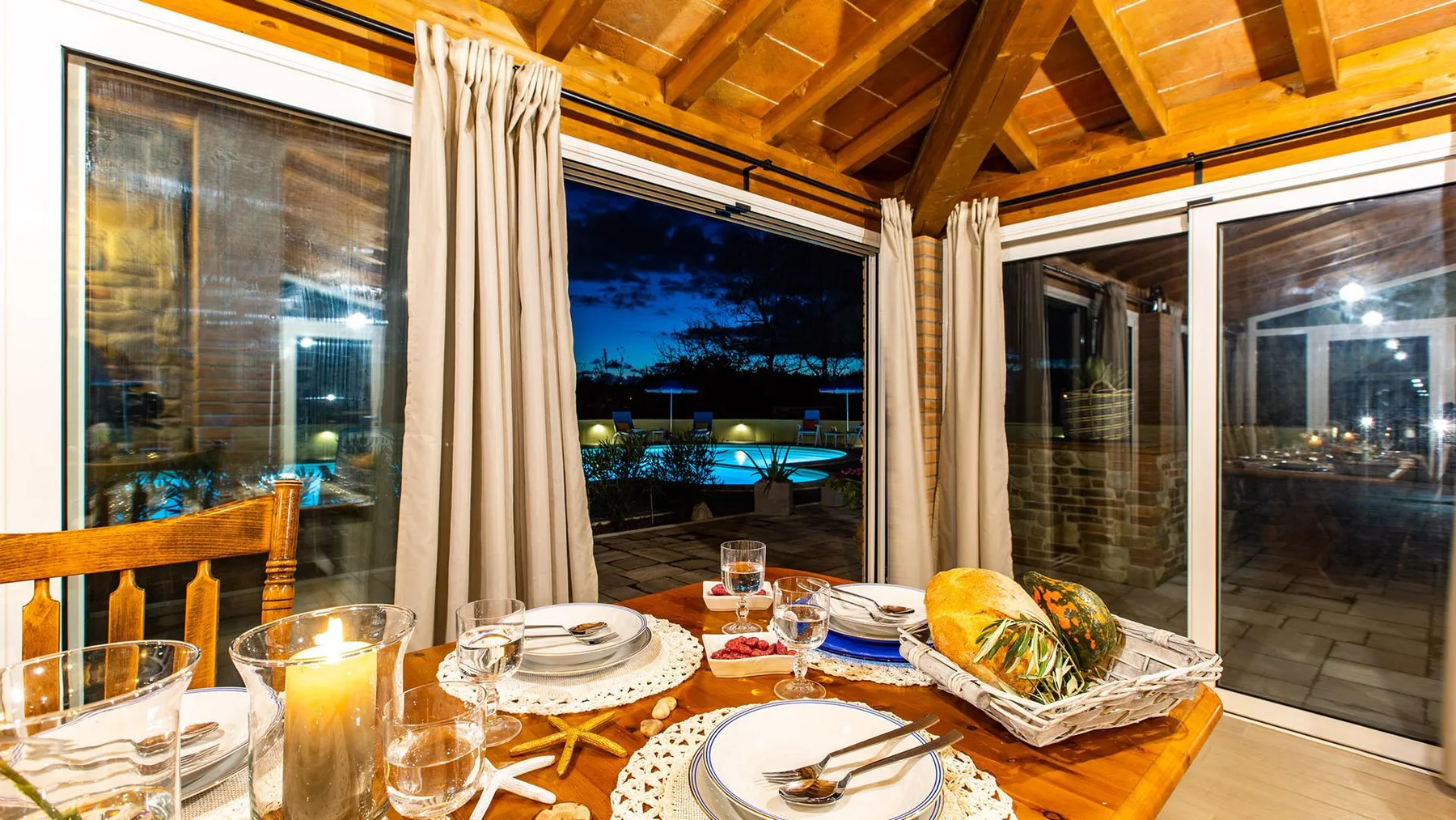 A dining table sits beside large windows overlooking an illuminated outdoor pool at night.