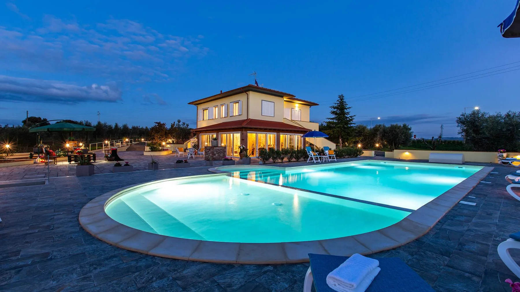 An illuminated outdoor swimming pool sits beside a two-story building at dusk.
