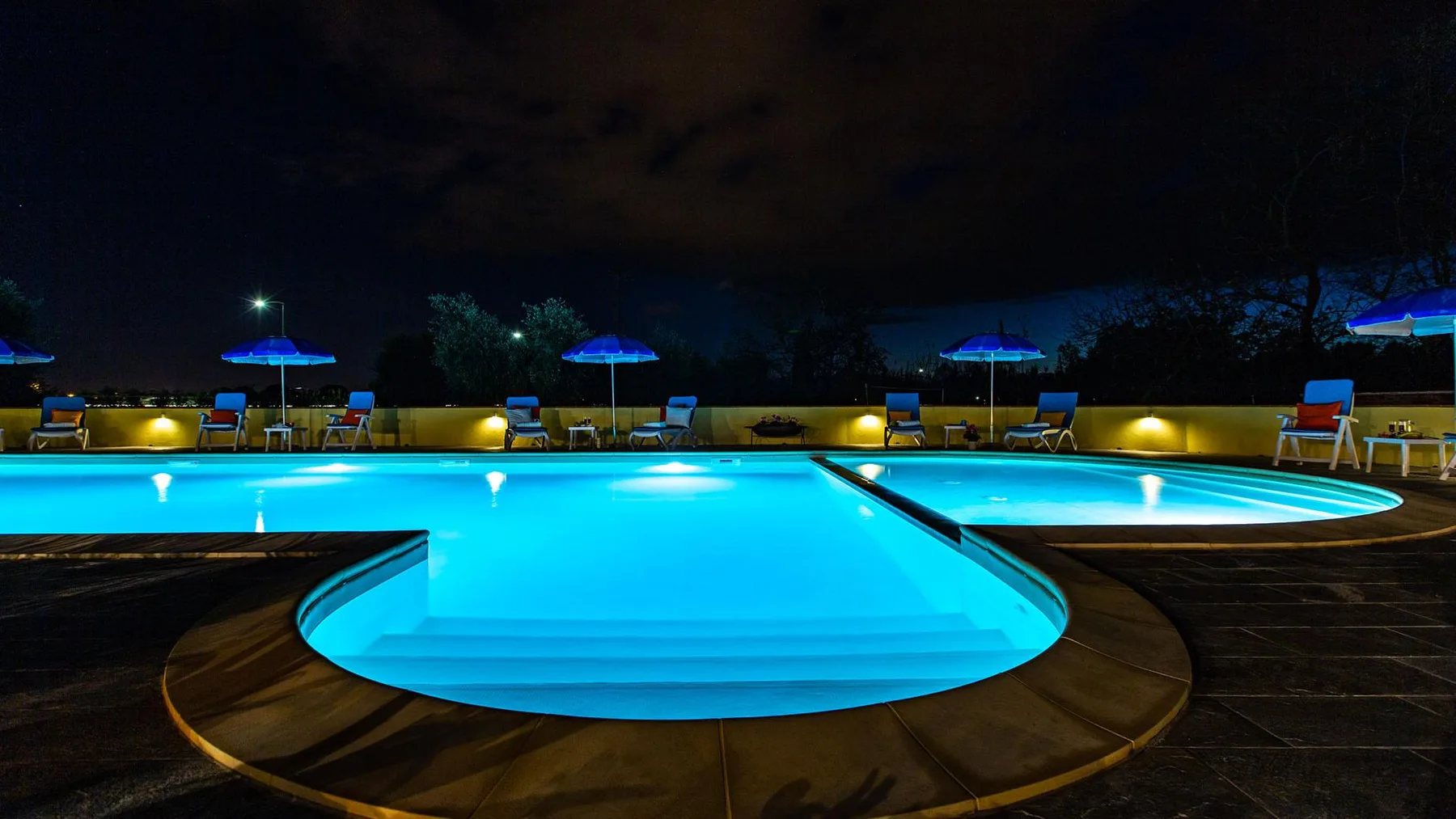 A brightly illuminated swimming pool at night, surrounded by lounge seating.