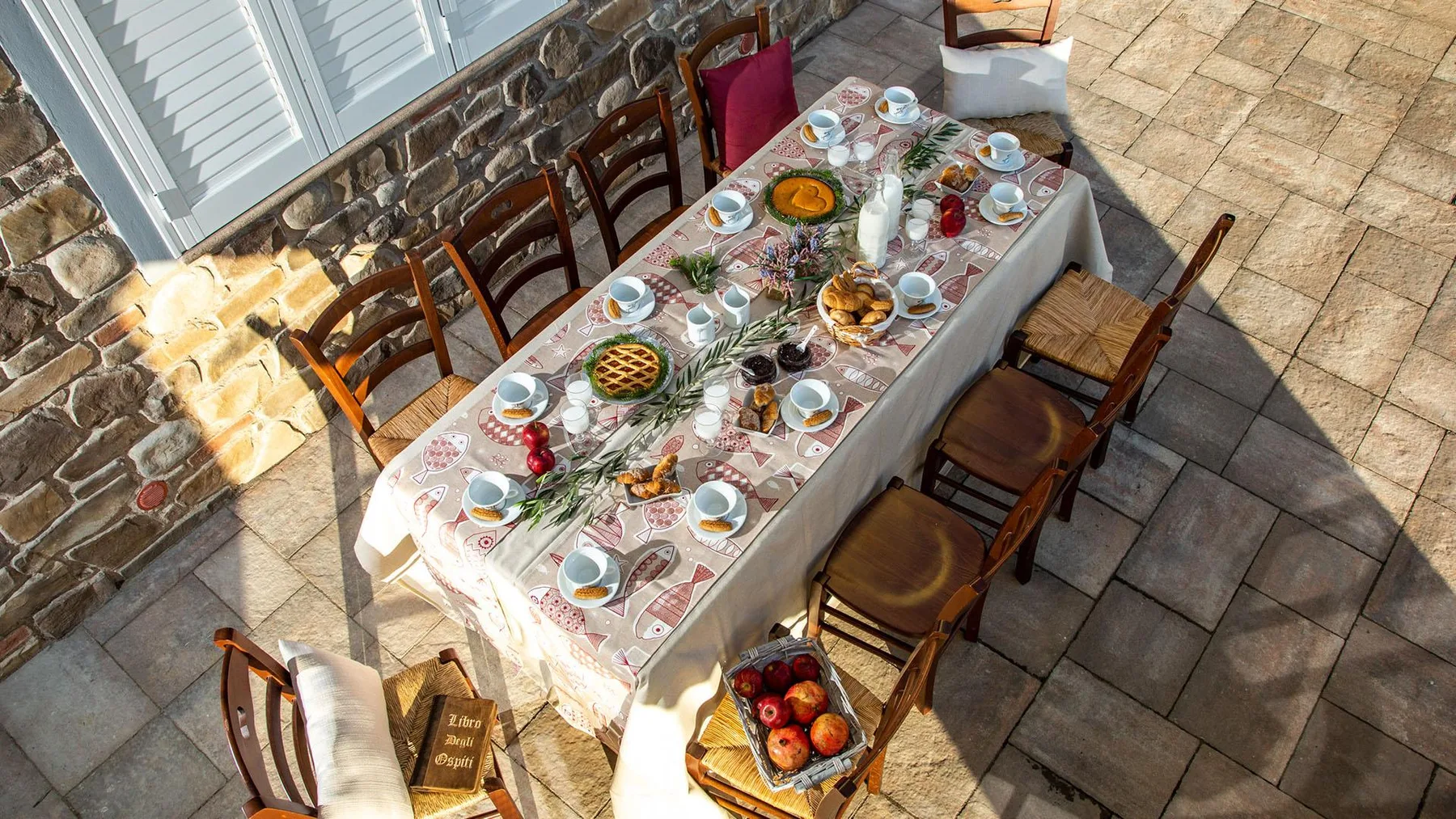 A long dining table is set with plates and food on a stone patio.