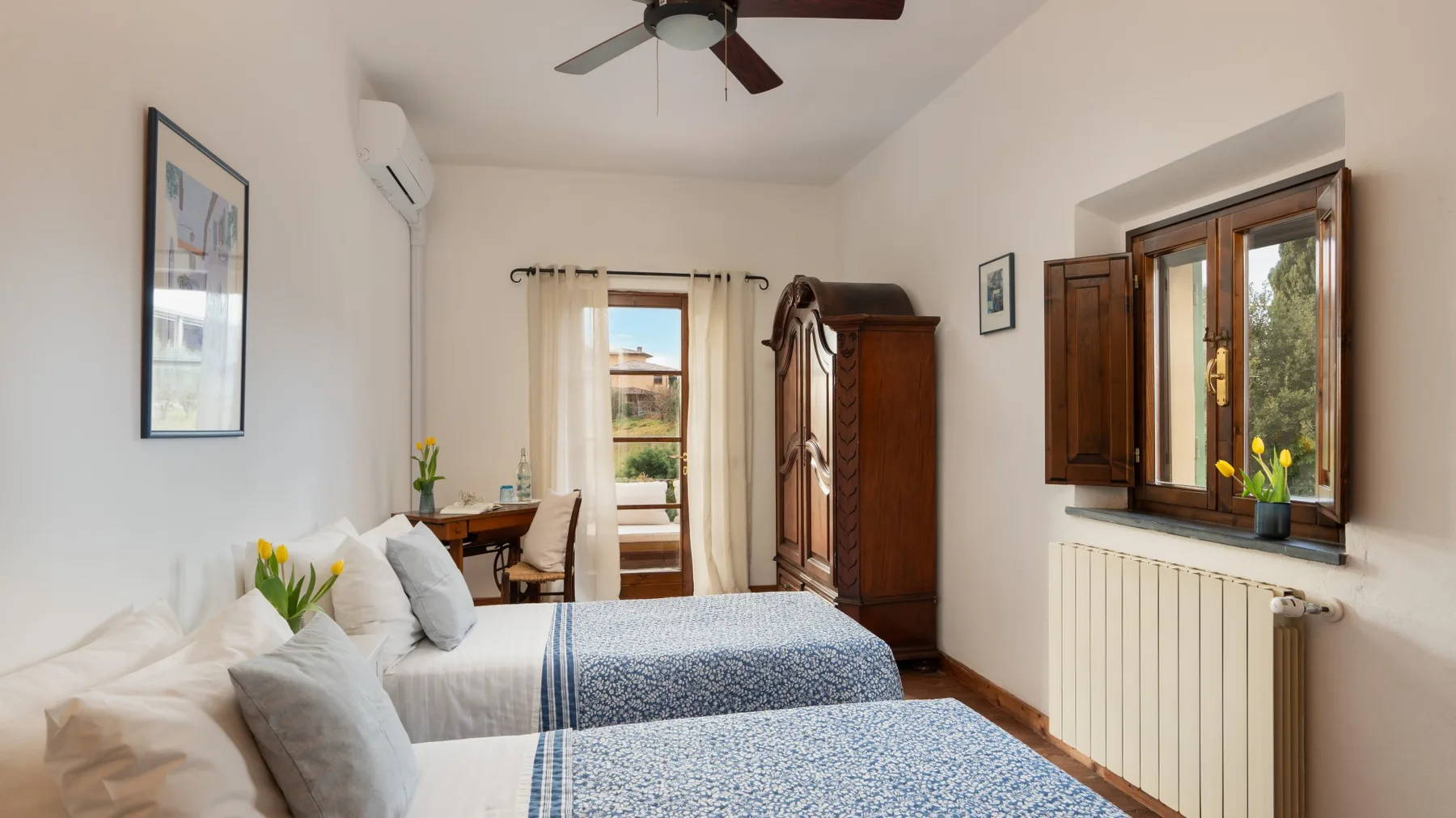 Un dormitorio luminoso con camas, un armario y ventanas con vistas a la vegetación.