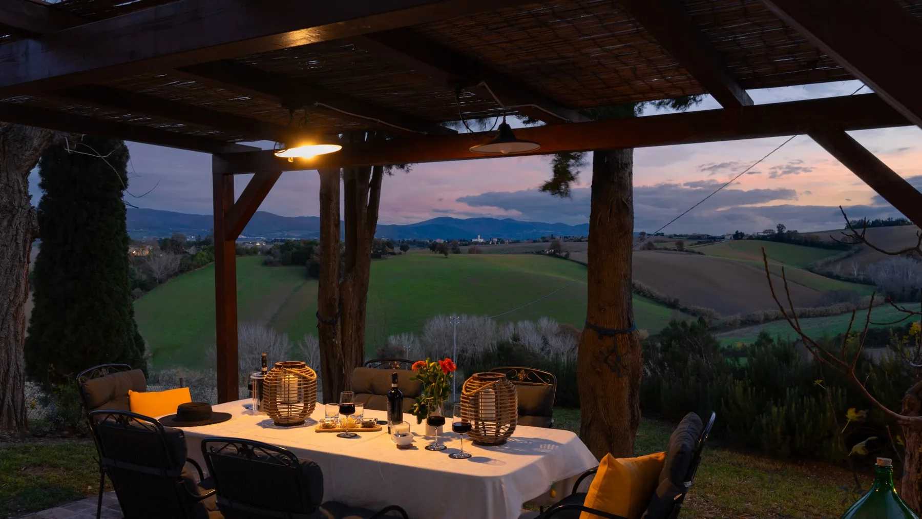 Una zona cubierta para comer al aire libre tiene vistas a campos y colinas al anochecer.