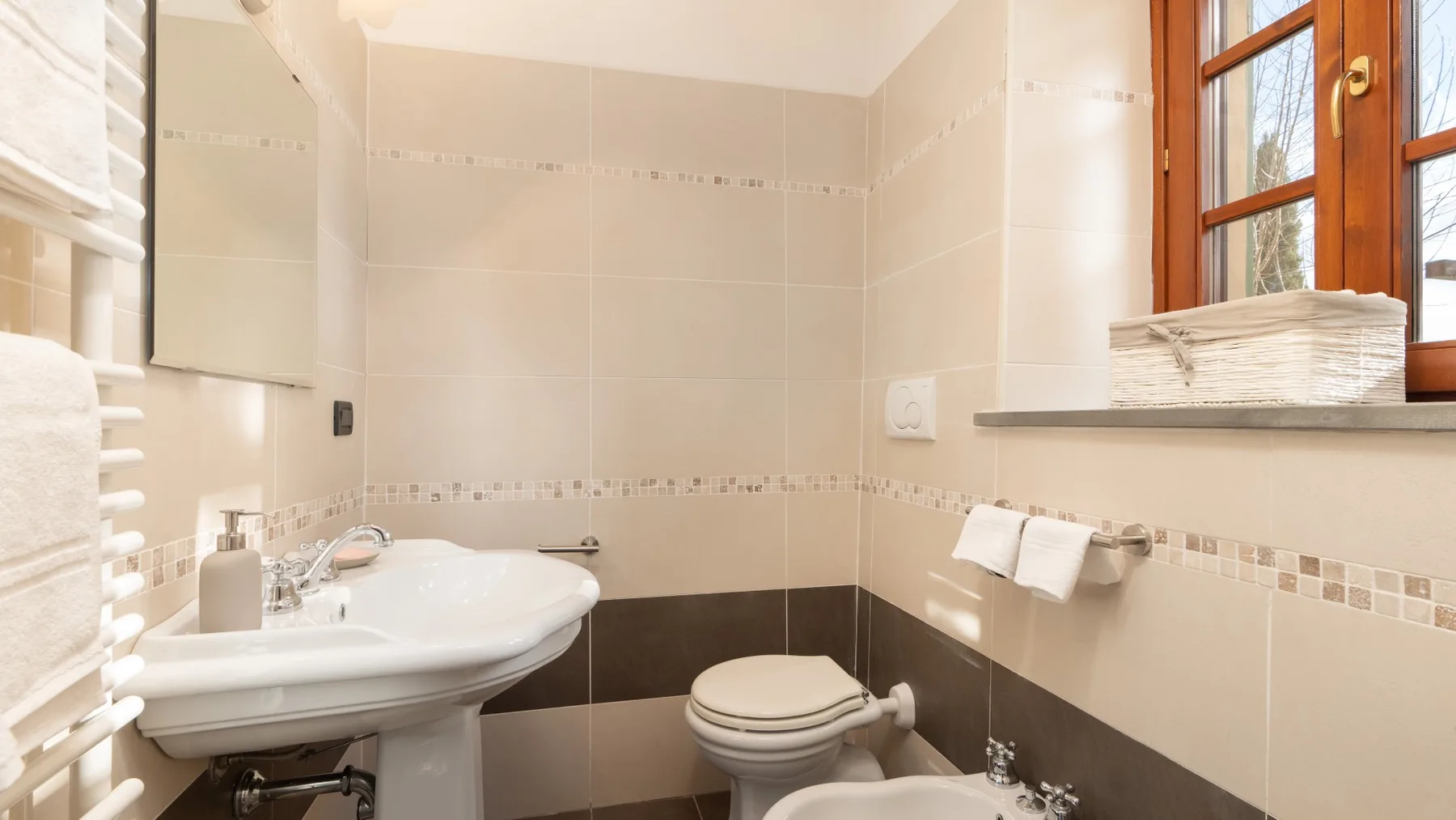 Un baño con azulejos, lavabo con pedestal e inodoro junto a una ventana.