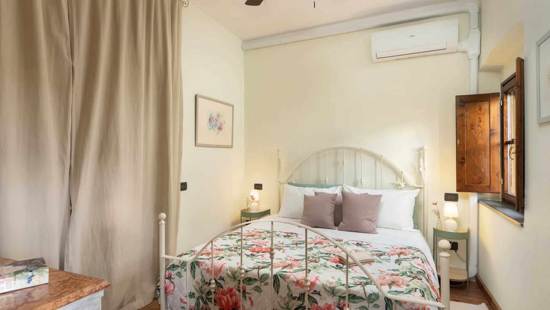 Un dormitorio con una colcha floral, mesitas de noche y una ventana.