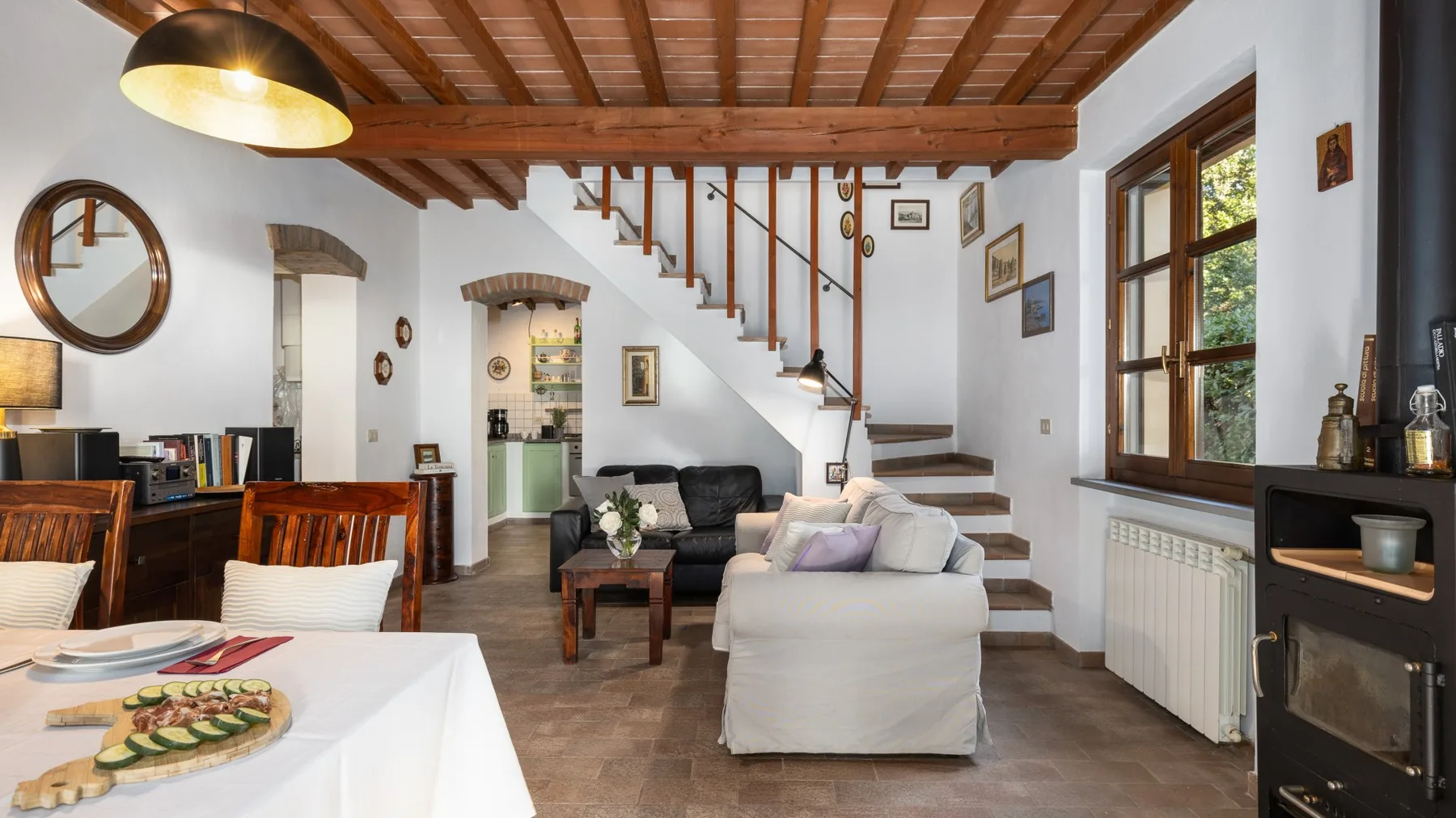 Una sala de estar con sofá, mesa de comedor y escalera bajo vigas de madera.