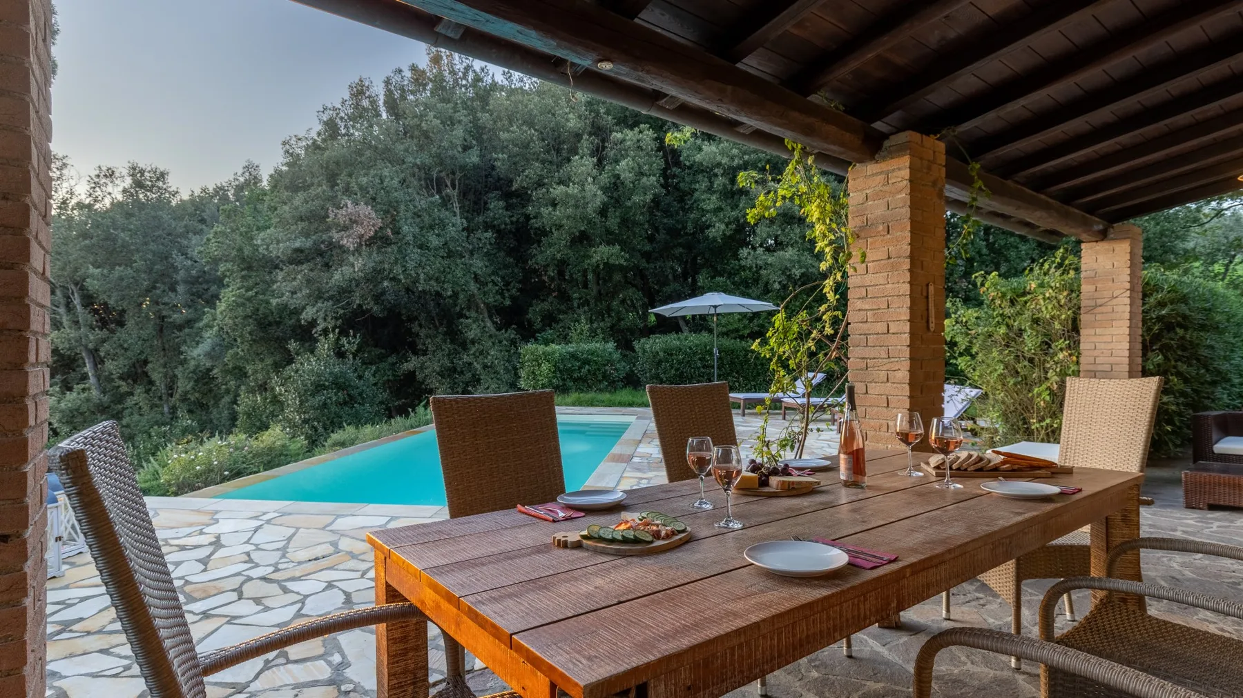 Un comedor exterior cubierto da a una piscina rodeada de árboles.