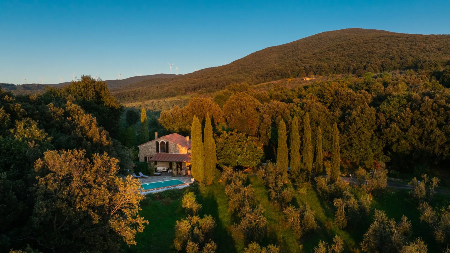 Una casa de campo y una piscina están entre árboles, bajo una colina boscosa.