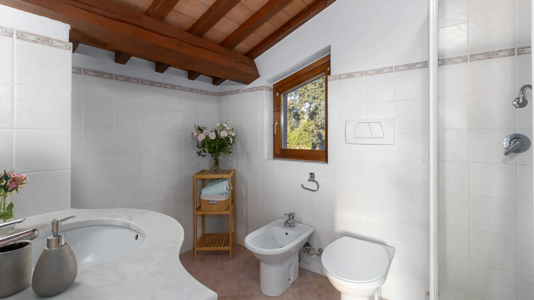 Un baño alicatado con lavabo, inodoro y una pequeña ventana bajo un techo de madera.