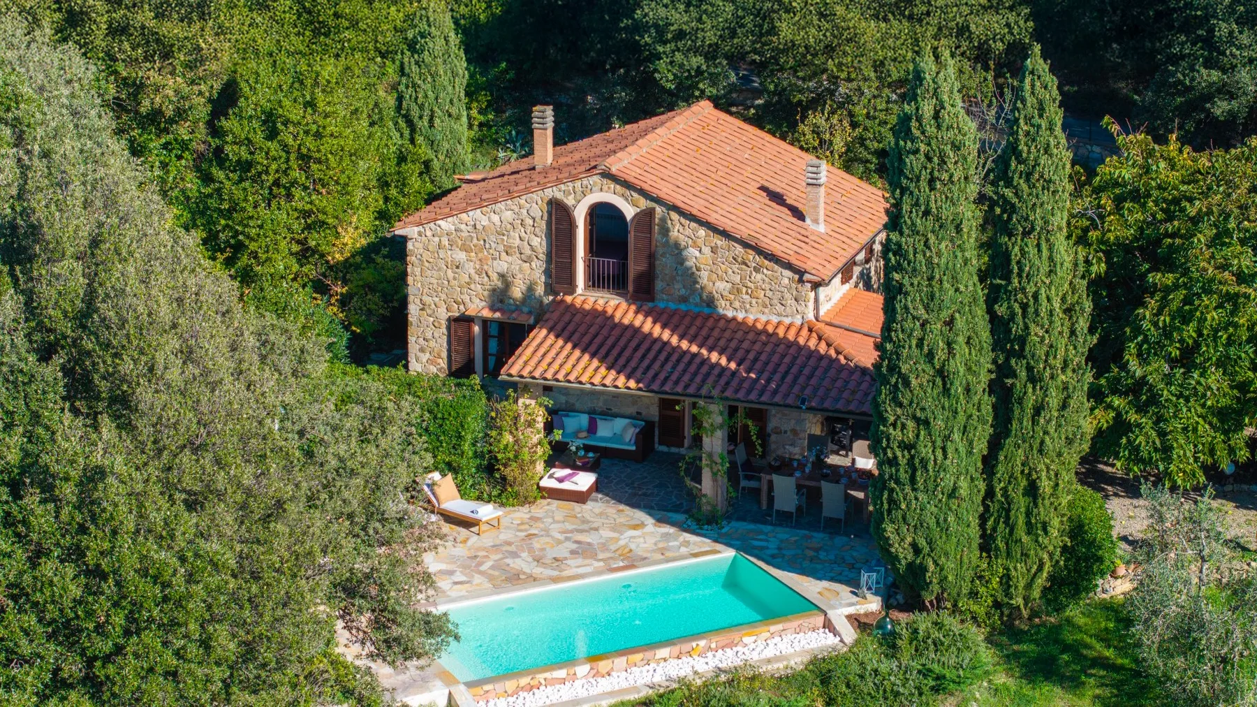 Una casa de piedra con techo de terracota está junto a una piscina turquesa, rodeada de árboles.
