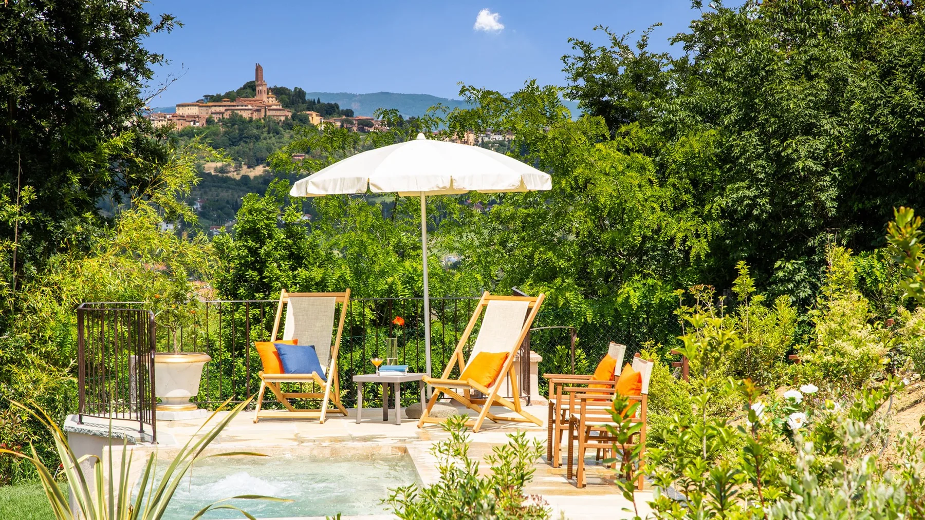 A garden patio with lounge seating, an umbrella, and a small pool overlooks a distant hillside town.