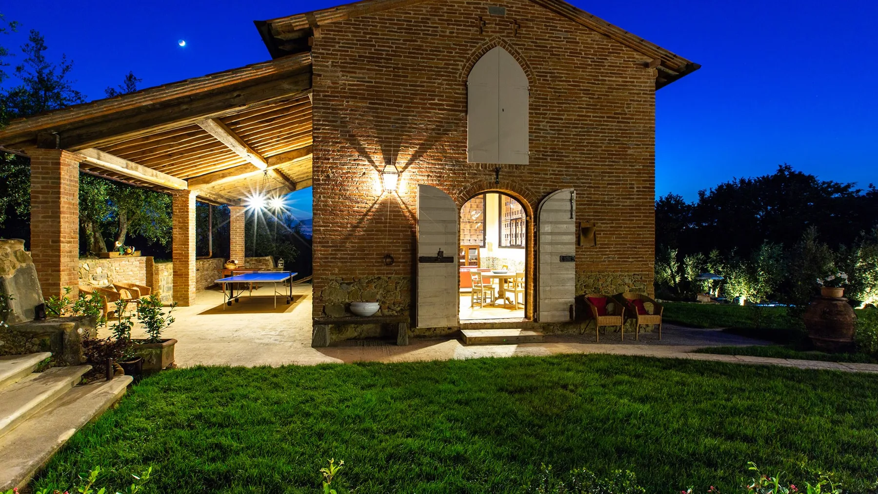A brick house with an illuminated covered patio opens onto a lawn at dusk.