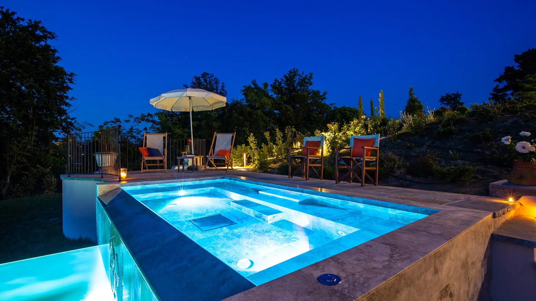An illuminated outdoor swimming pool sits beside seating in a landscaped garden at dusk.