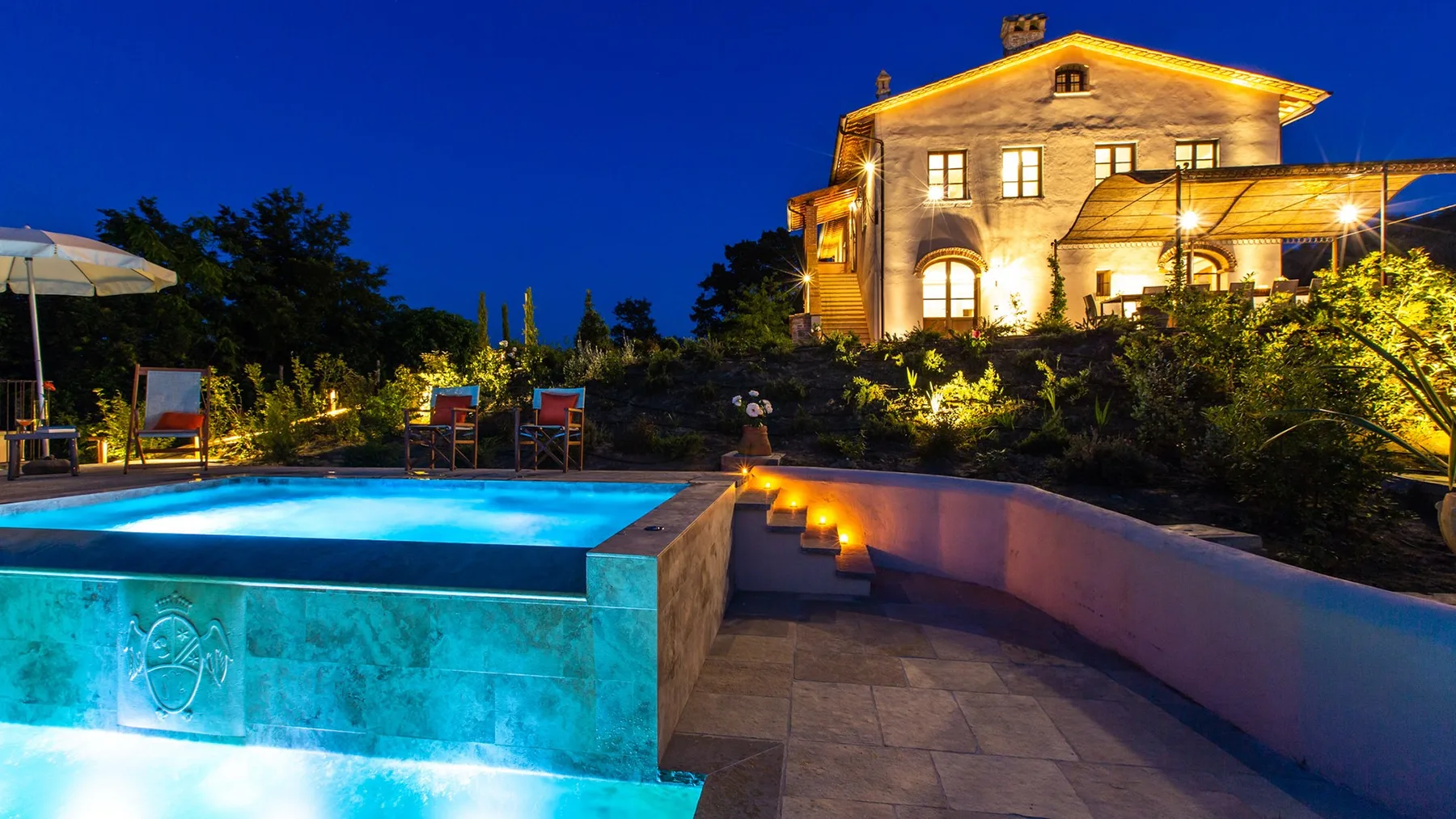 A lit house overlooks a swimming pool and curved stone patio at dusk.