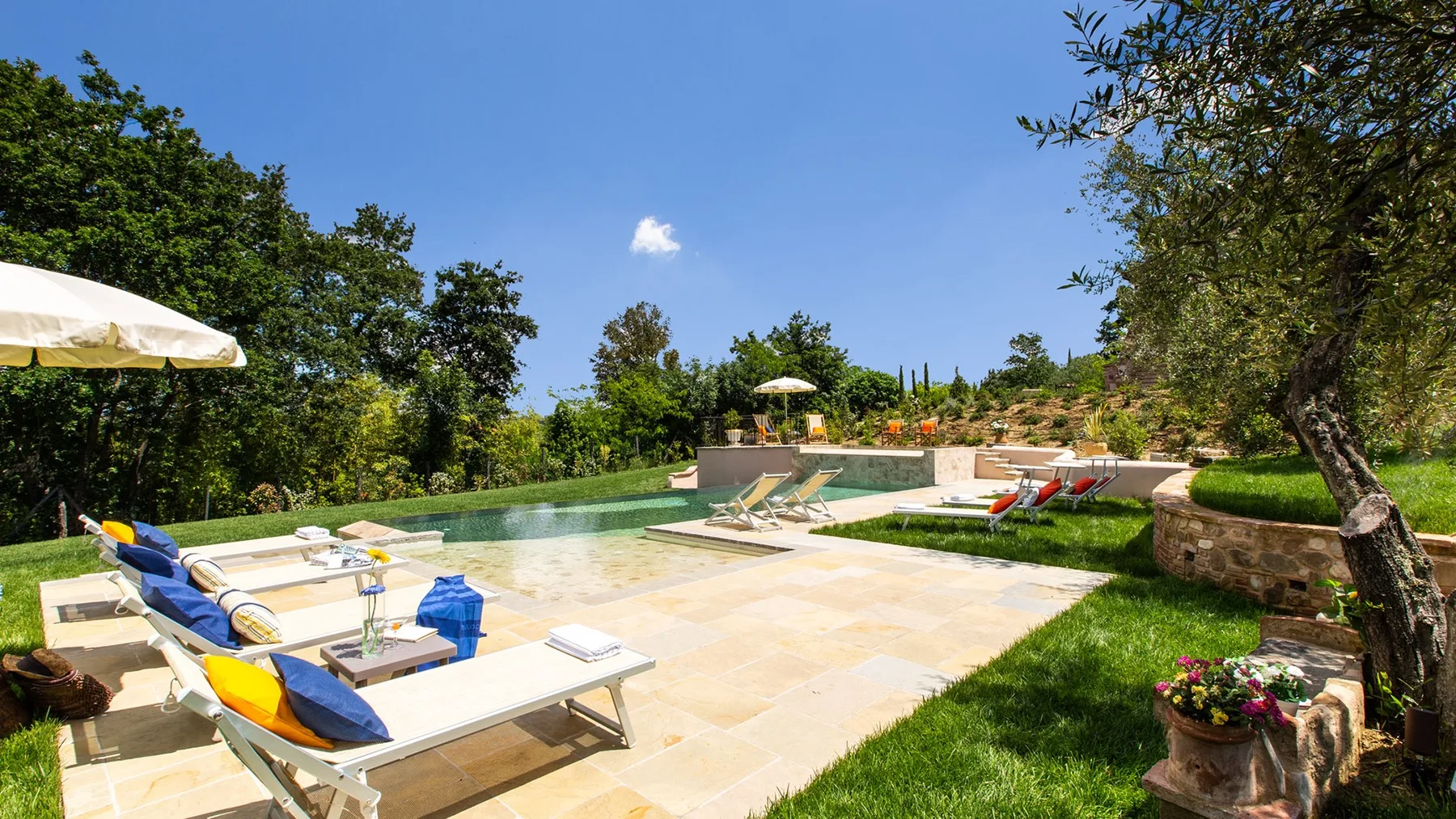 An outdoor swimming pool sits beside a pale stone terrace with sun loungers and greenery.