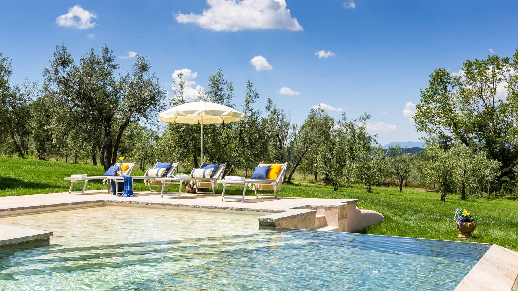 An outdoor swimming pool beside a grassy garden with loungers and an umbrella.