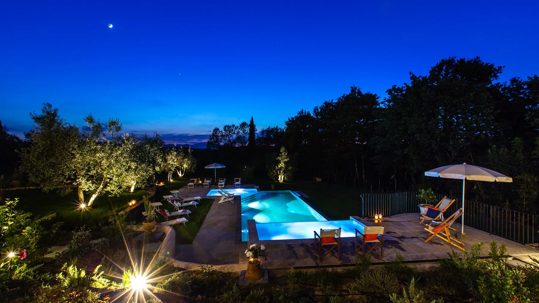 A swimming pool glows at dusk beside a landscaped garden and outdoor seating.