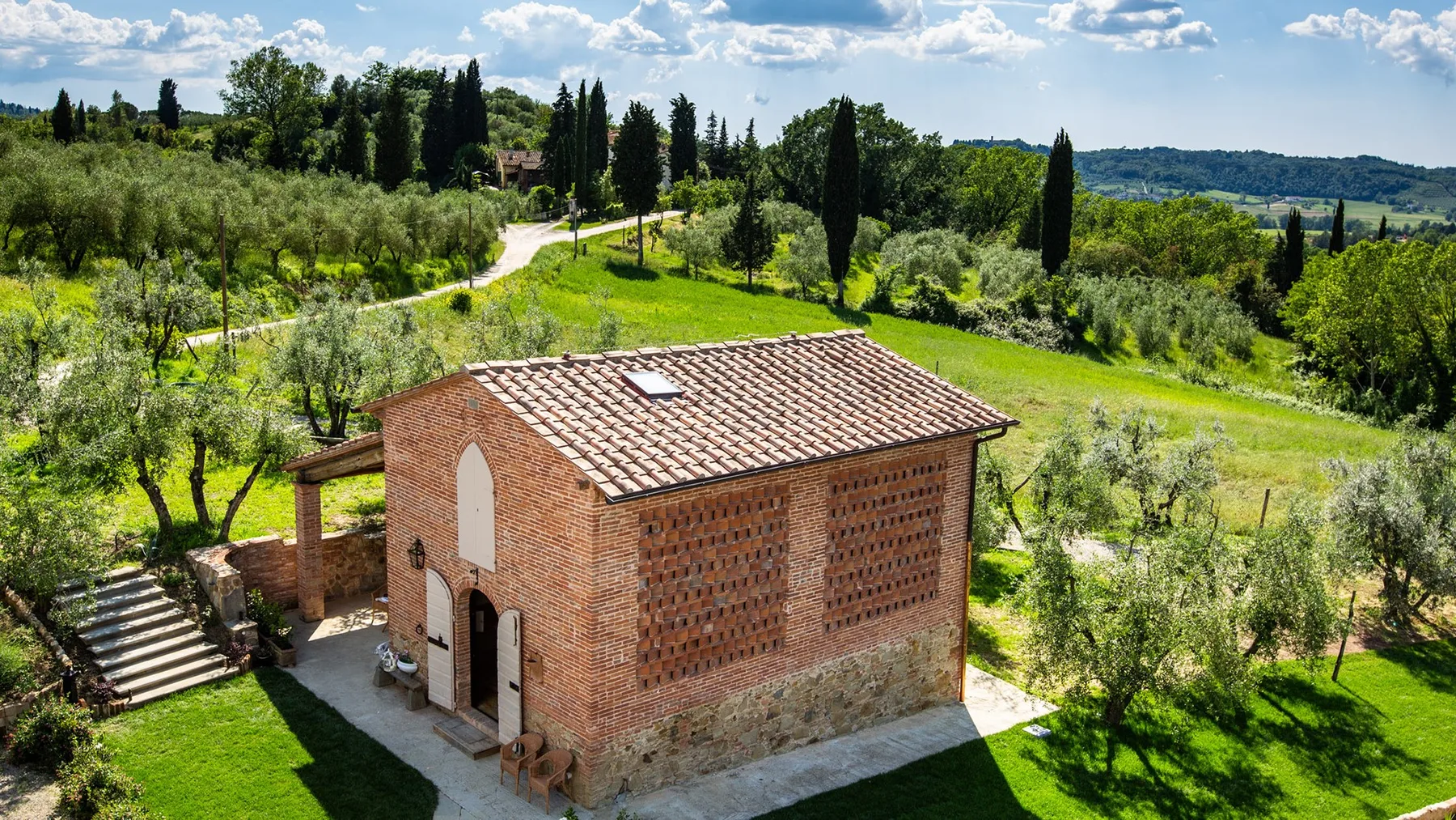 A brick house with a tiled roof sits amid green fields and trees.