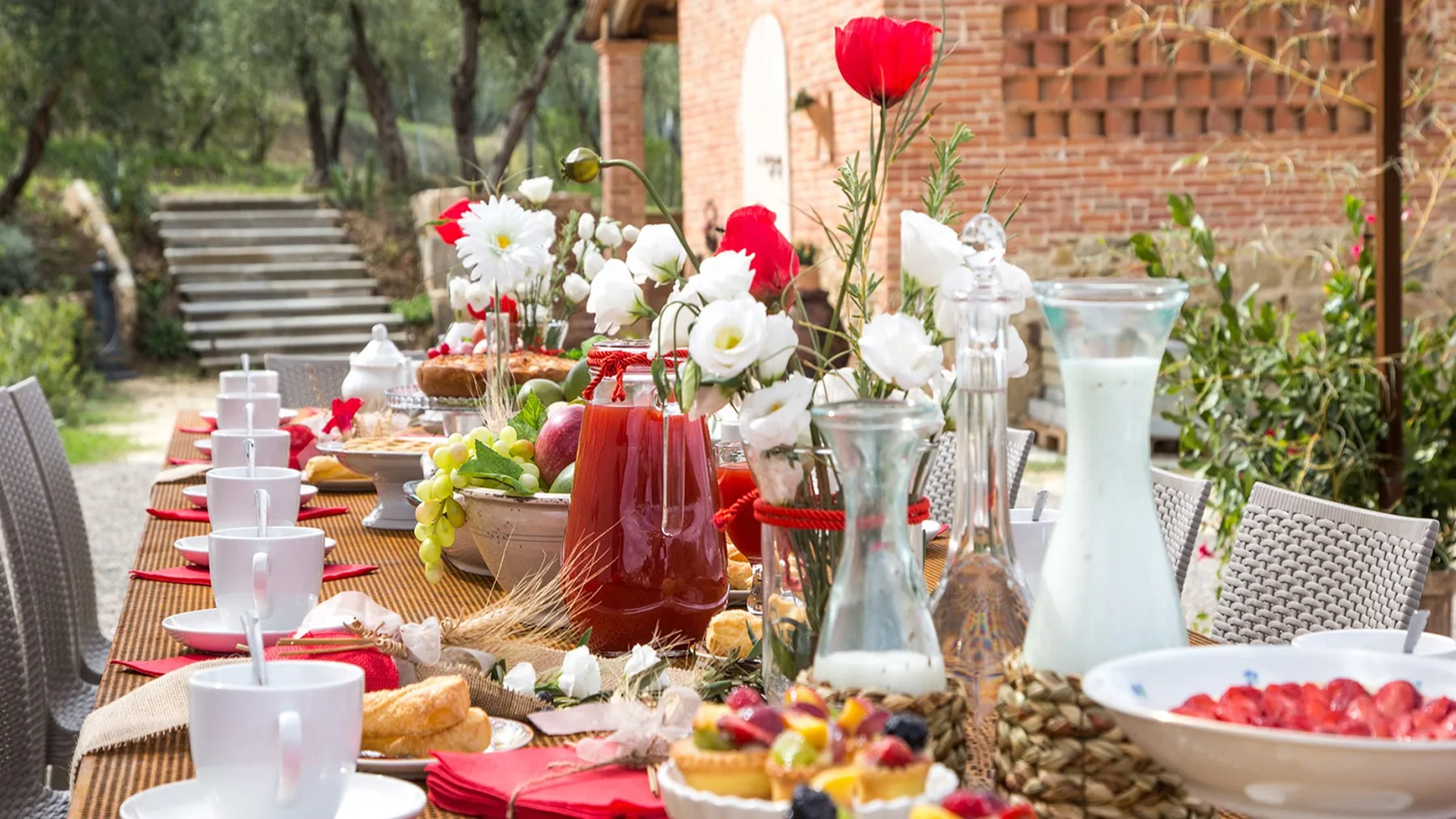 An outdoor dining table is set with dishes, drinks, and flowers beside a brick wall.