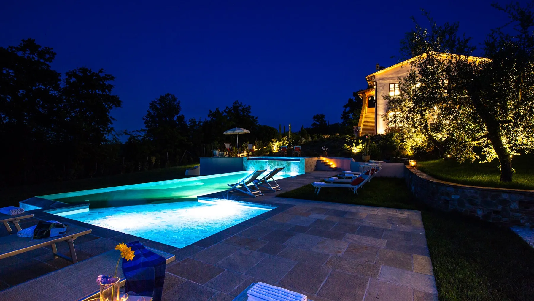 A swimming pool and paved patio are illuminated beside a building at night.