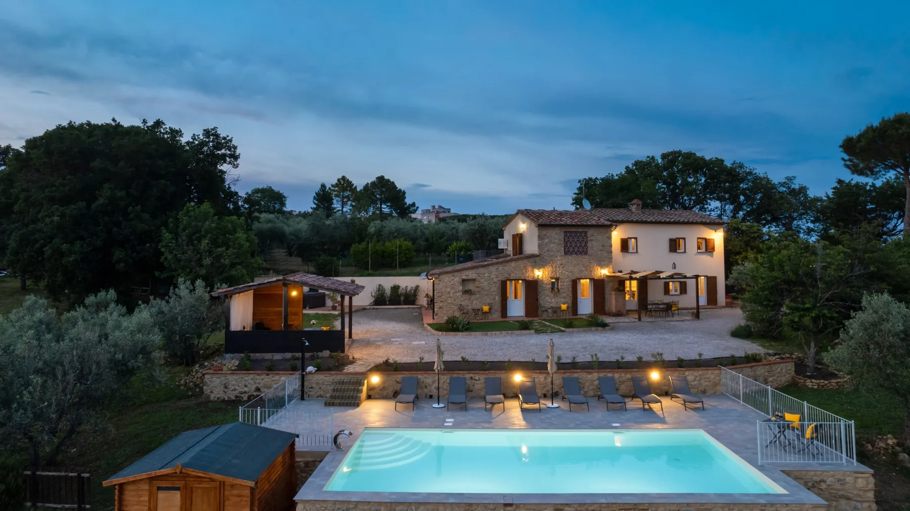 A country house at dusk overlooks a lit swimming pool and outdoor seating.