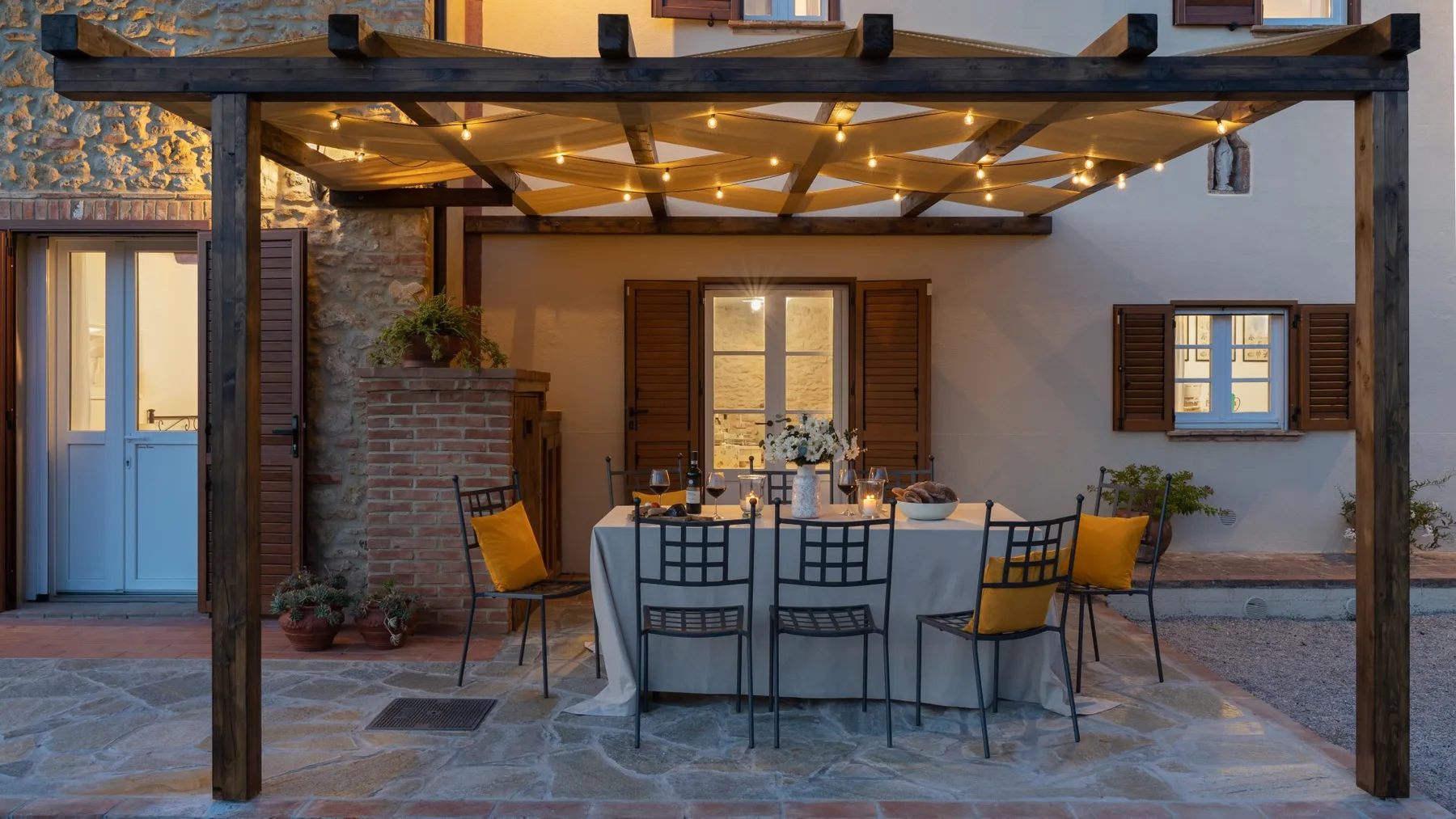 An outdoor dining area beneath a wooden pergola, with a set table and surrounding seating.
