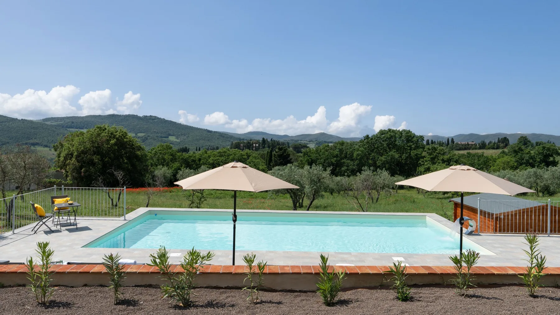 Outdoor swimming pool with patio umbrellas, greenery, and hills in the distance.