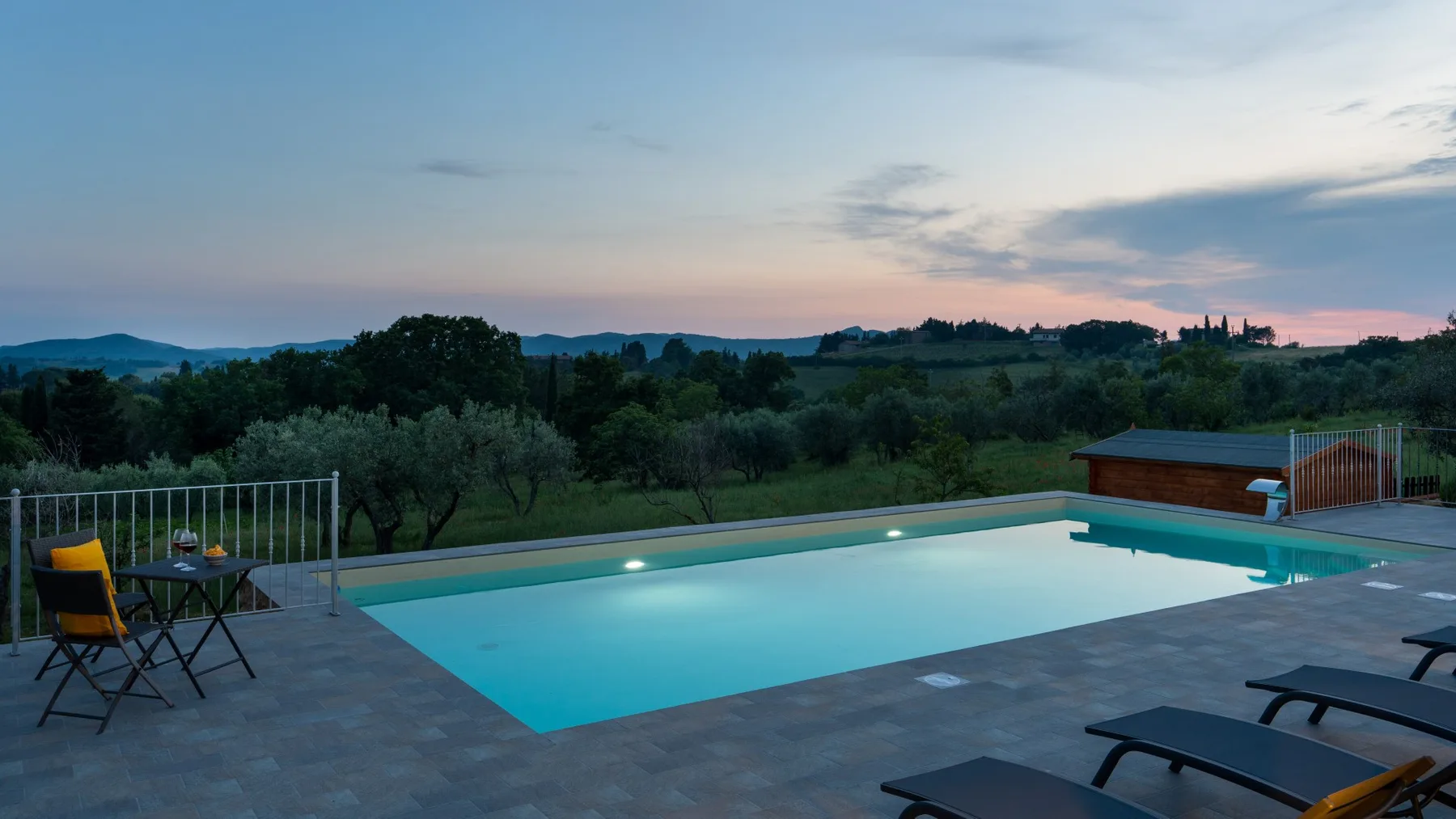 An outdoor swimming pool beside a patio, with trees and hills in the background.