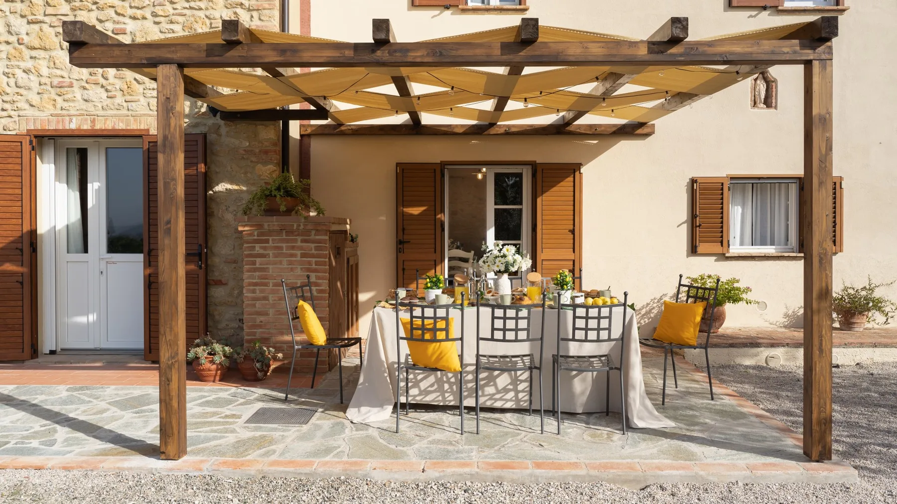 An outdoor dining area sits beneath a wooden pergola beside a stucco building.