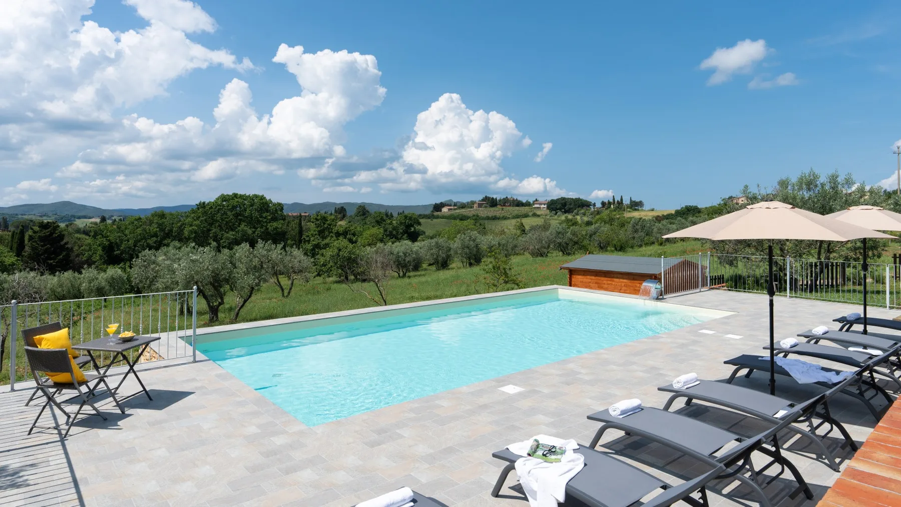 An outdoor swimming pool and surrounding patio overlook green hills beneath a blue sky.