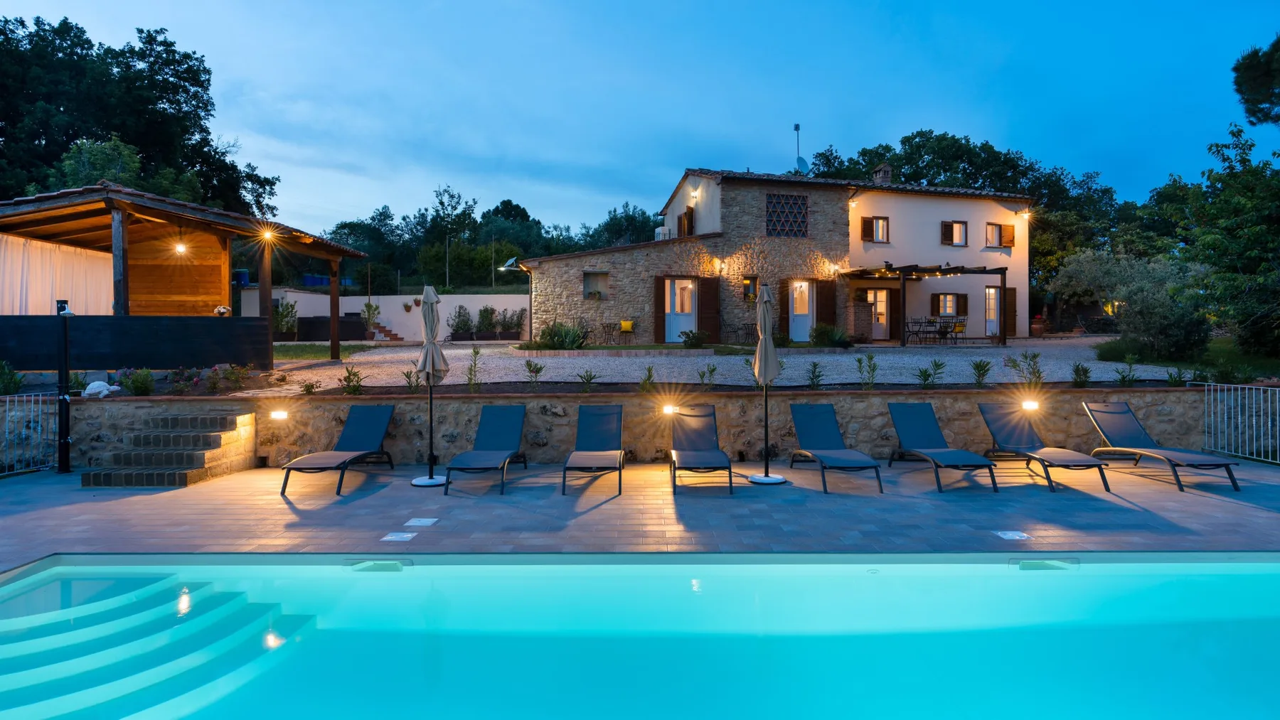 An outdoor swimming pool and lounge seating sit in front of a lit stone house at dusk.