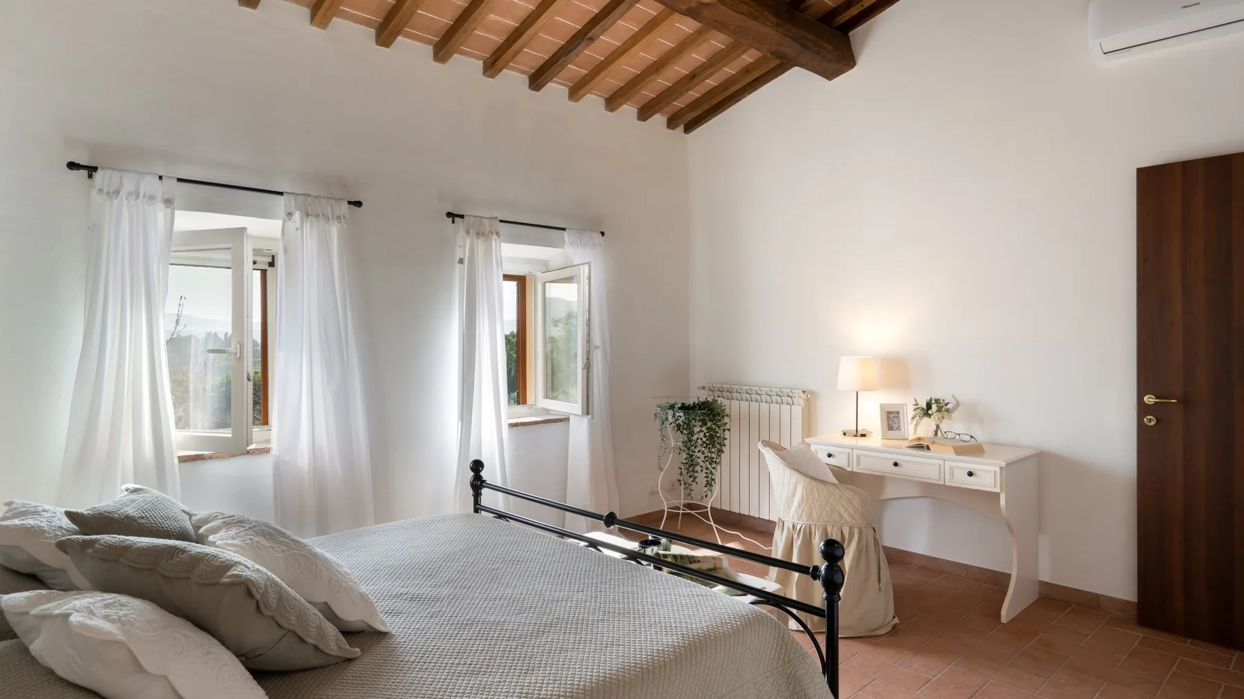 A bedroom with a bed, two curtained windows, and a desk beneath a wooden-beamed ceiling.
