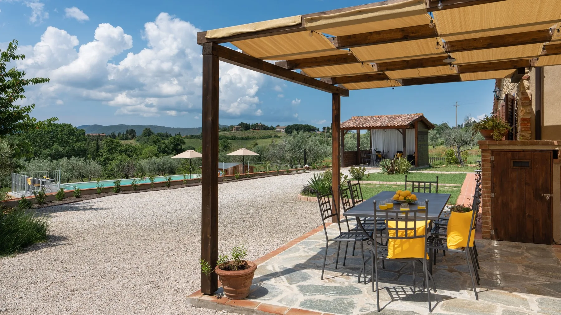 An outdoor dining area sits beneath a wooden pergola beside a pool and green hills.