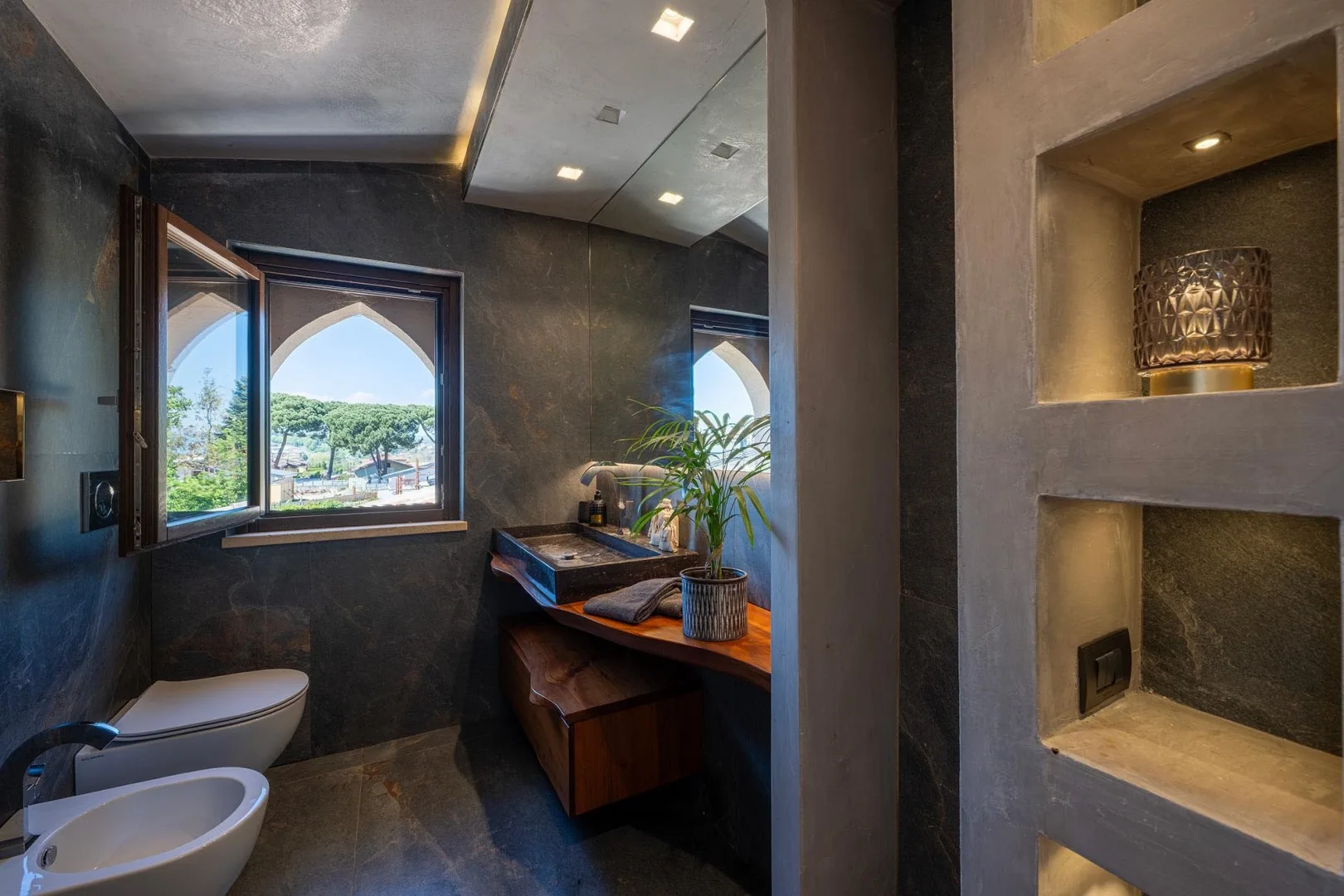 A bathroom with a vanity, toilet, and windows overlooking greenery.