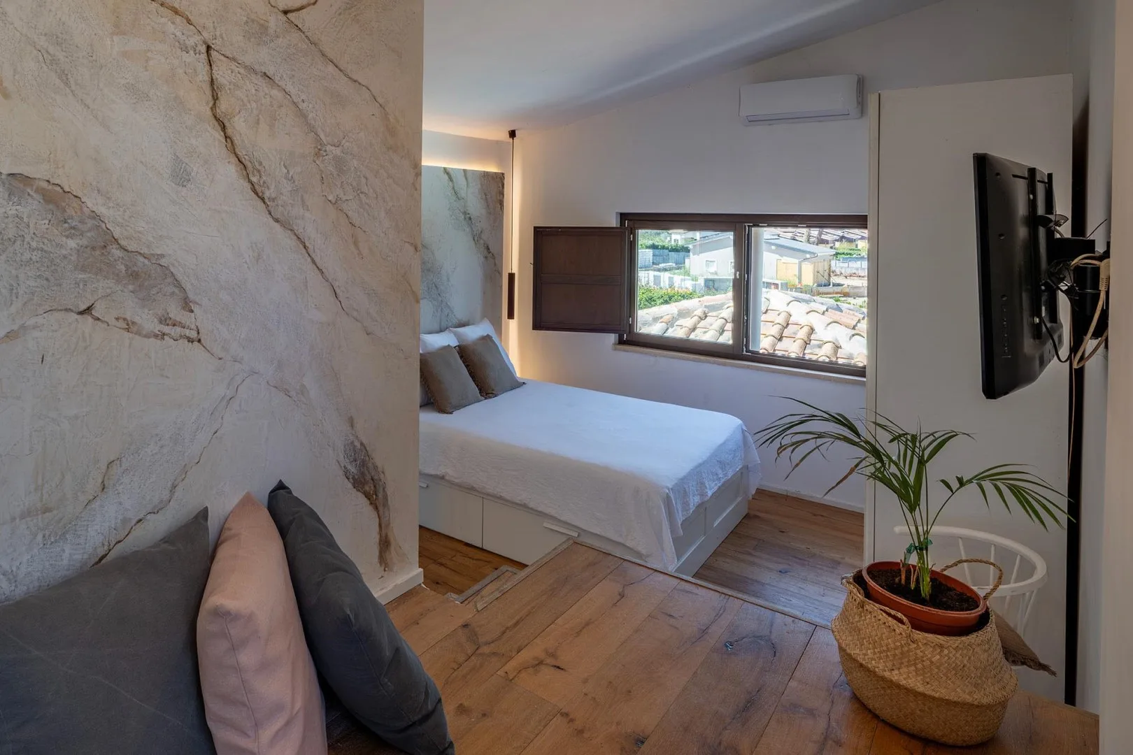A bedroom with a neatly made bed, a wide window, and a wall-mounted television.