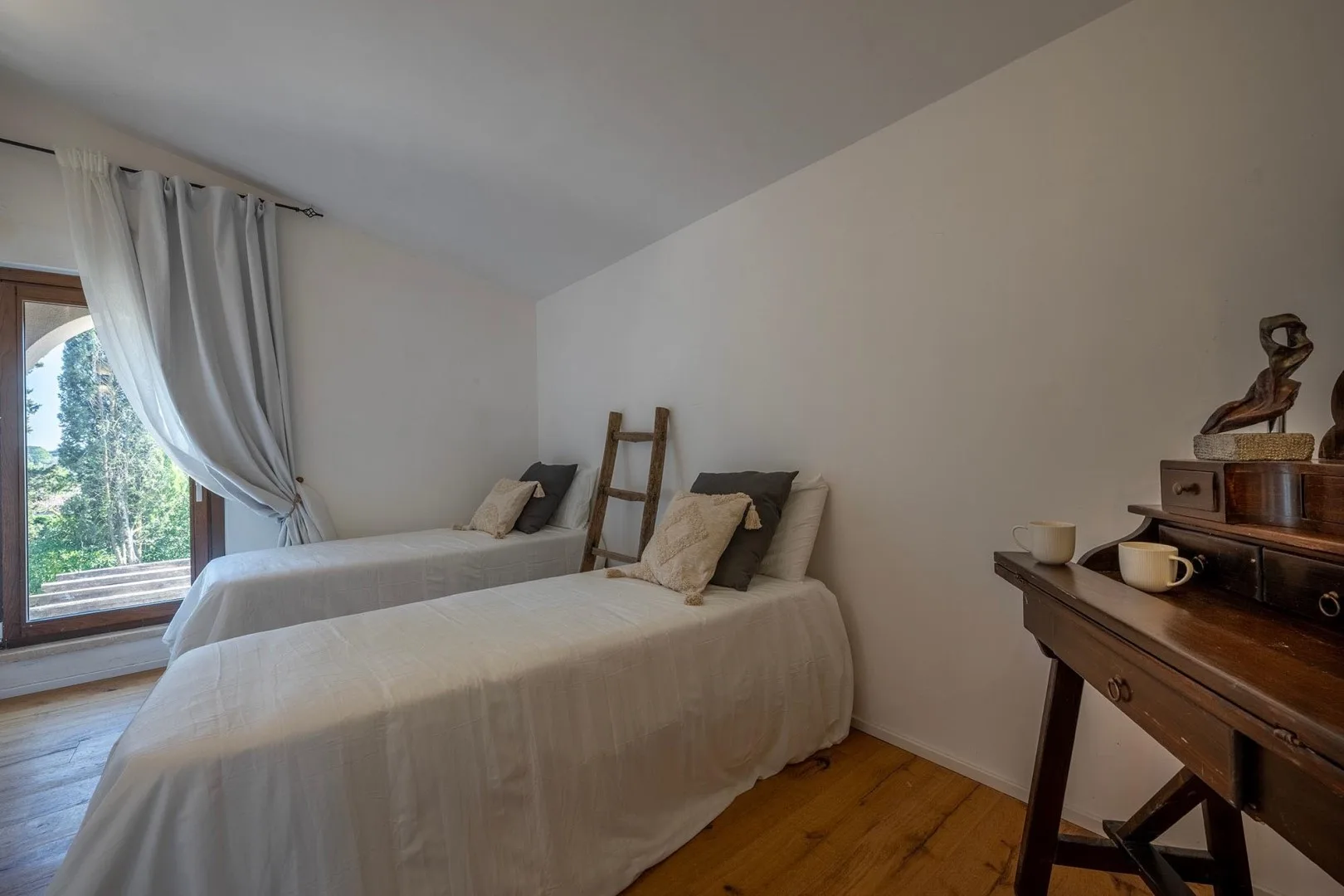 A bedroom with two single beds, light curtains, and wooden flooring.