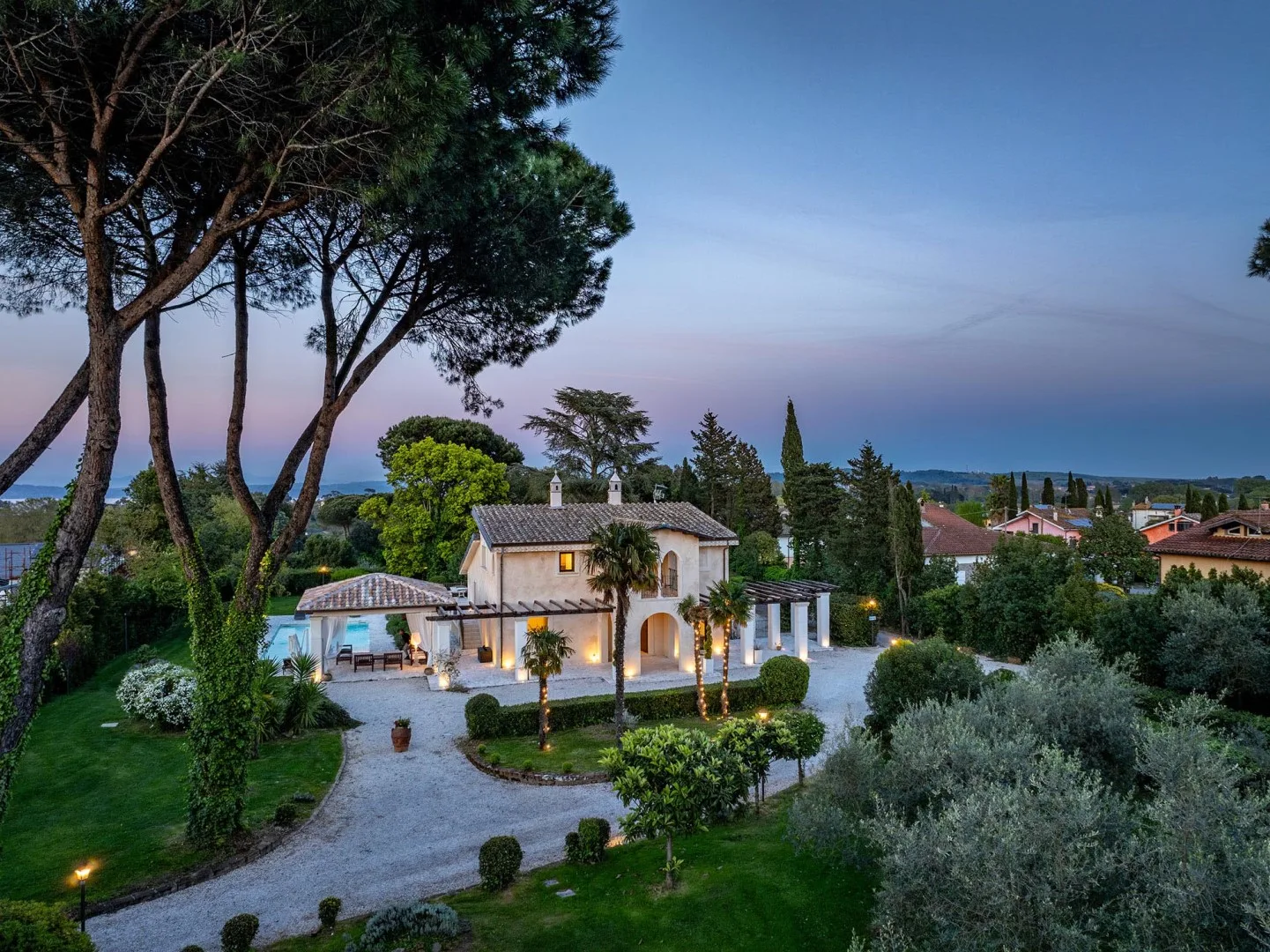A landscaped villa sits among trees, gardens, and a curved driveway at dusk.
