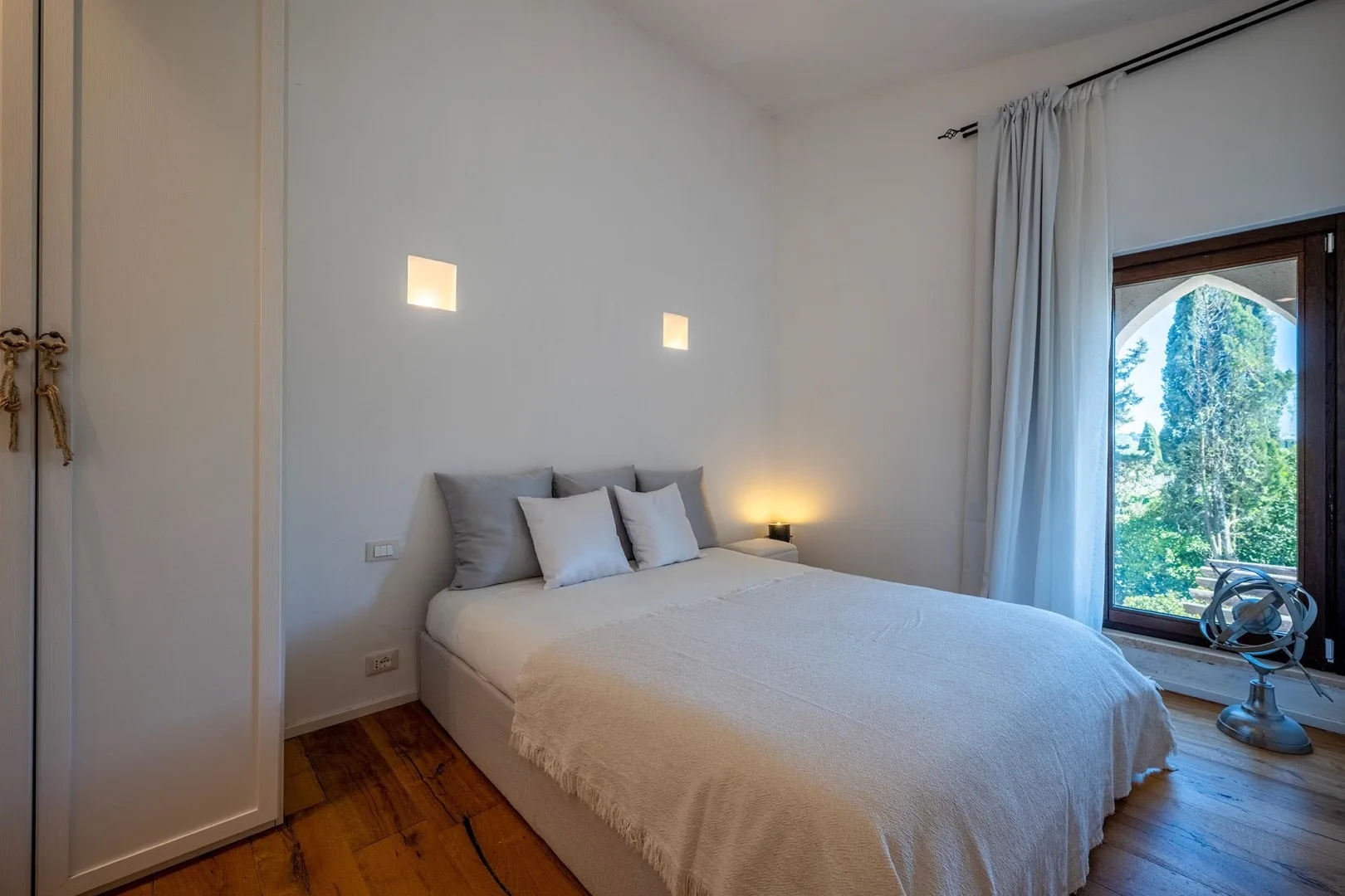 A bedroom with a bed beside a window overlooking greenery.