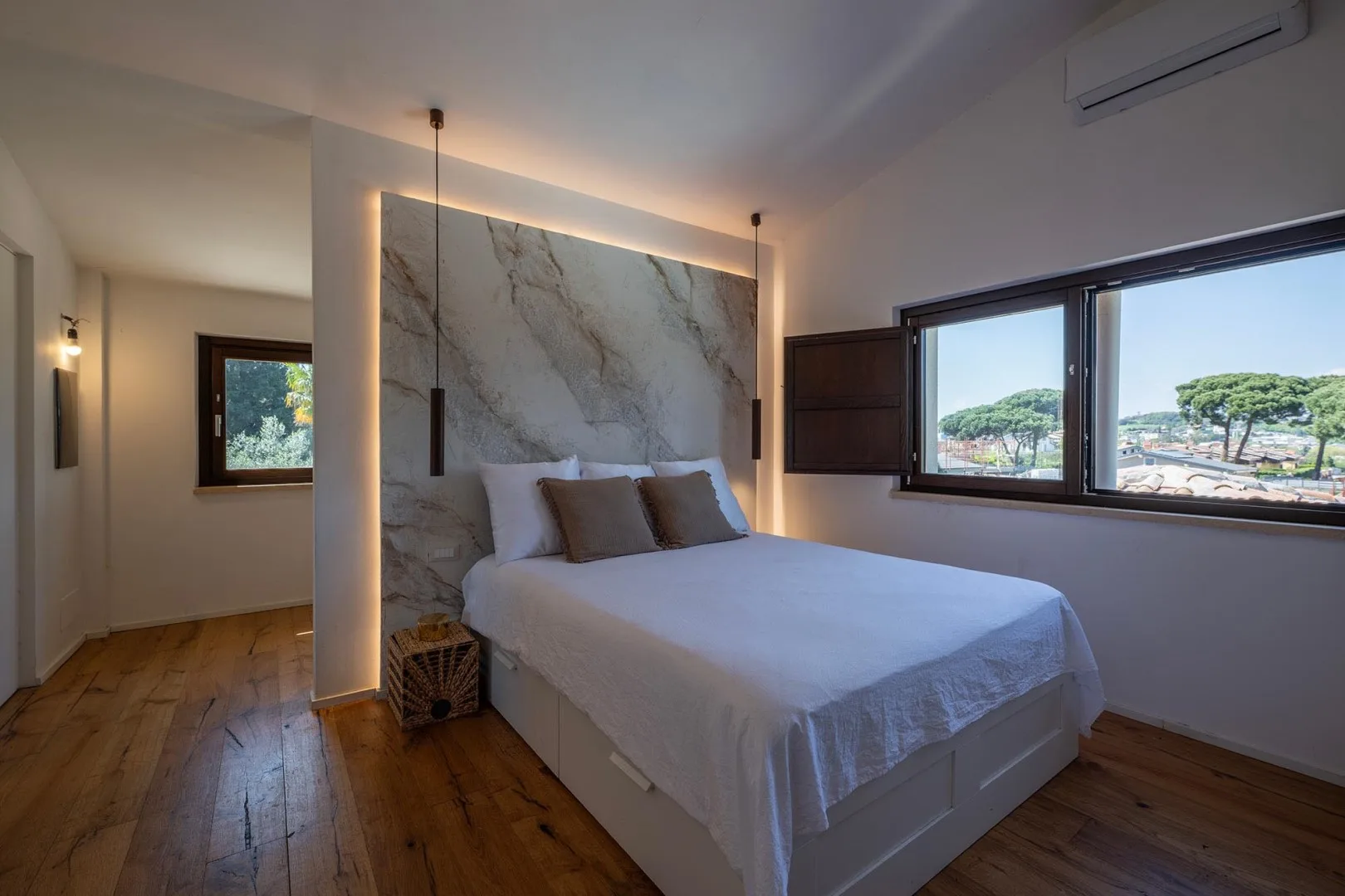 A bedroom with a bed against a marble-patterned wall and windows overlooking greenery.